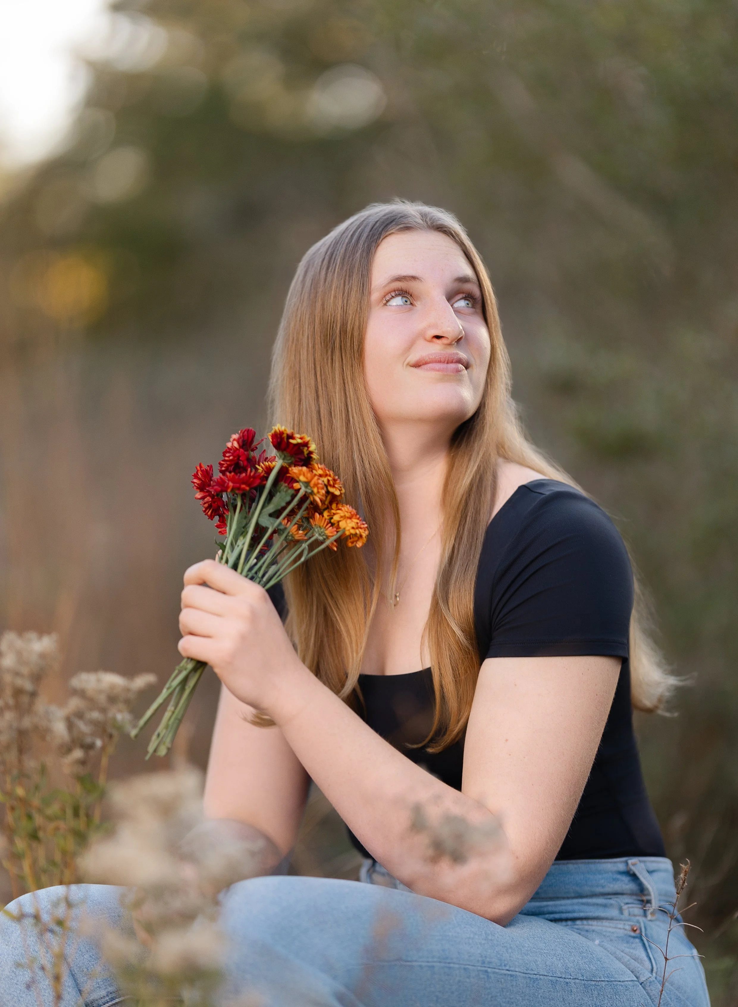 A young woman with long red hair sitting outdoors in a natural setting, holding a small bouquet of red and orange flowers, looking up with a thoughtful expression.