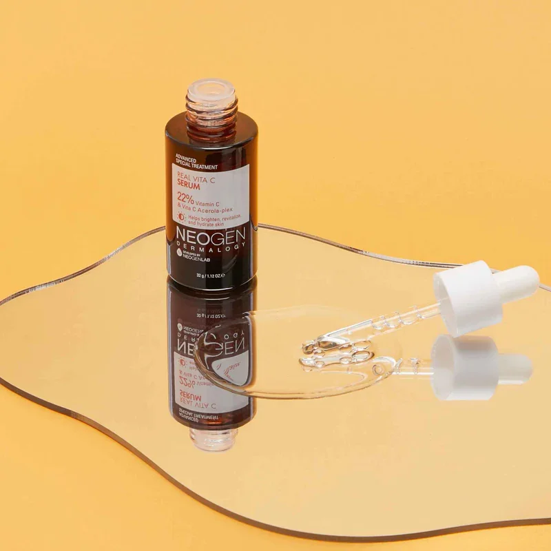 A brown bottle of NEOGEN Real Vita C Serum with a white dropper cap, placed on a reflective surface with a yellow background. The dropper contains clear serum, some of which is spilled on the surface.