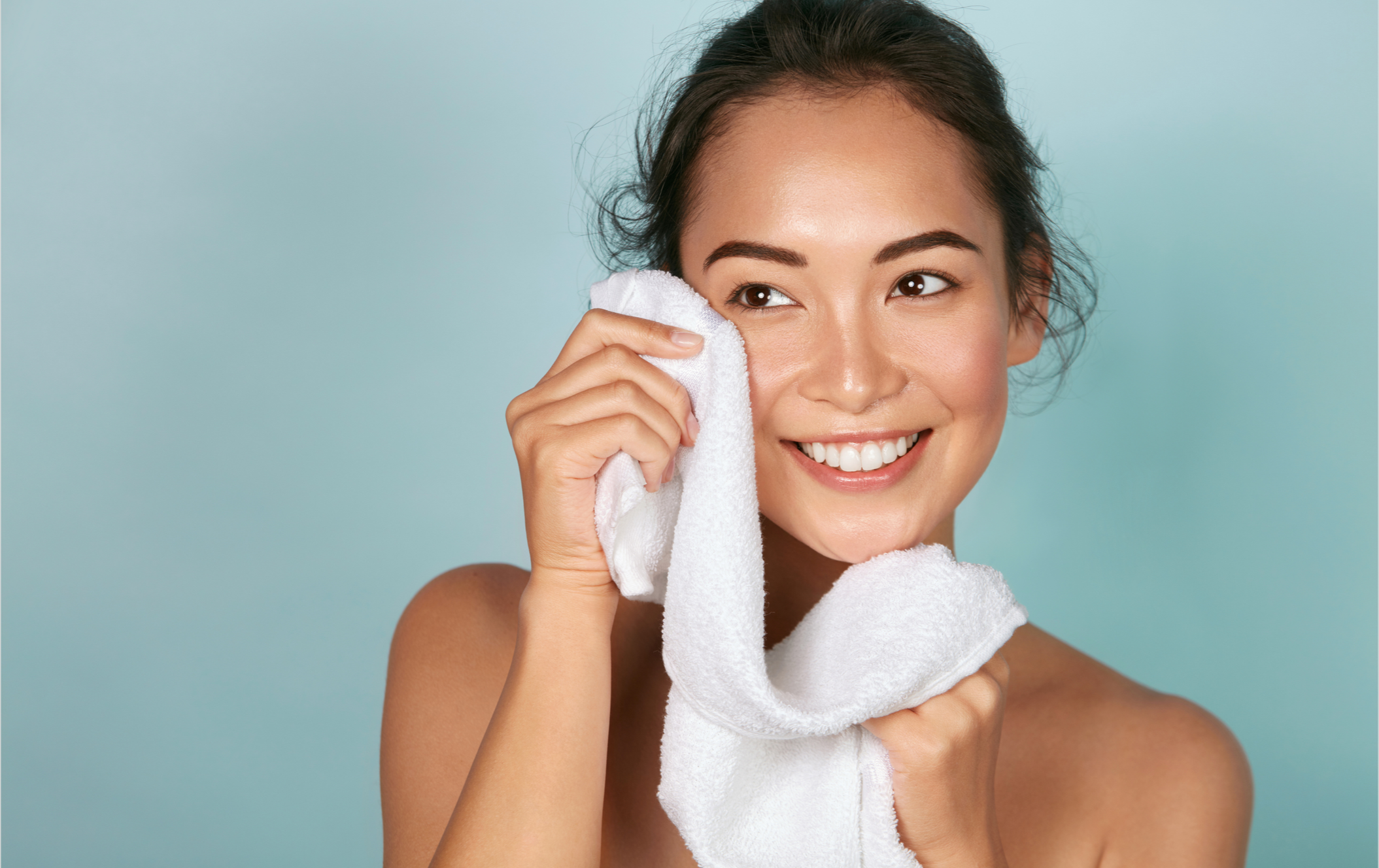 A woman smiling while holding a white towel to her face against a light blue background.