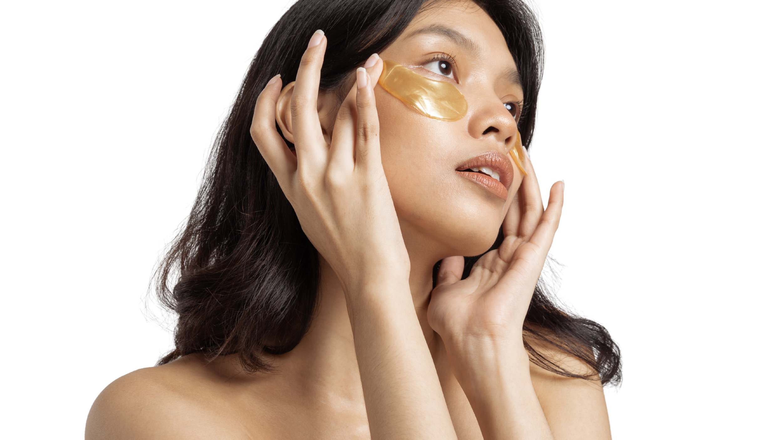 Woman with long dark hair applying a gold facial mask to her face, looking slightly upward.