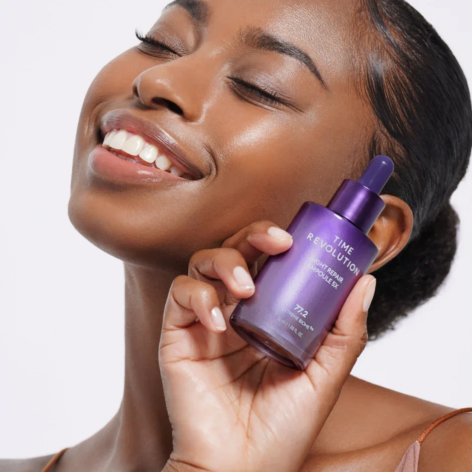 A woman with smooth skin smiling and holding a purple bottle of Time Revolution Night Repair Ampoule 5X close to her face.