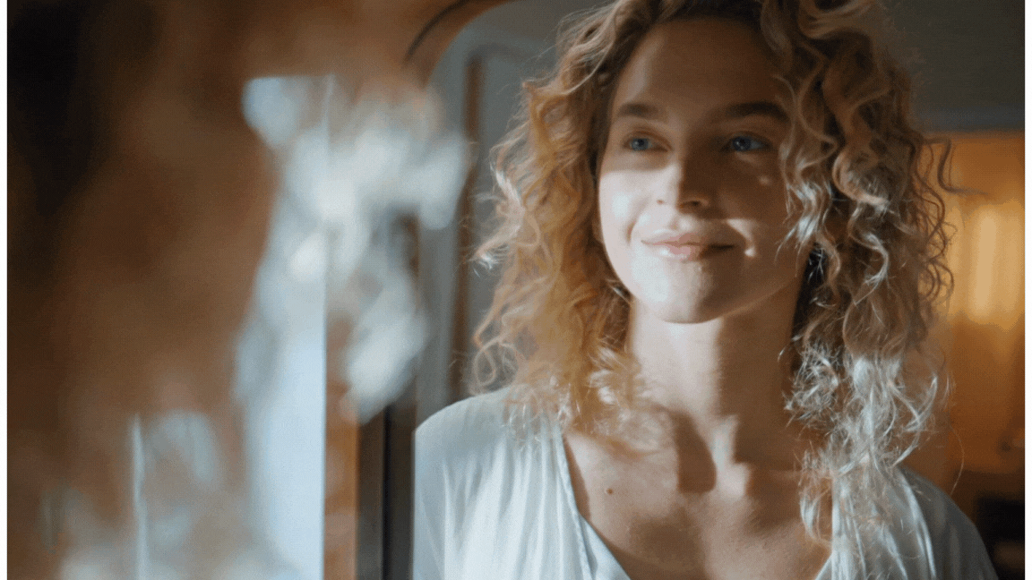 A woman with curly blonde hair smiling softly, facing another person who is blurred in the foreground.