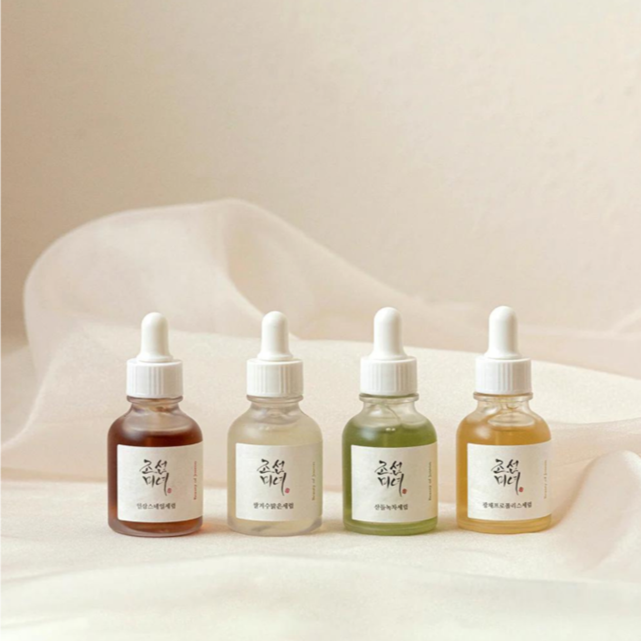 A collection of Beauty of Joseon skincare products, including four small glass bottles with droppers containing different colored liquids and a squeeze tube, arranged on a white surface with wooden decor pieces.