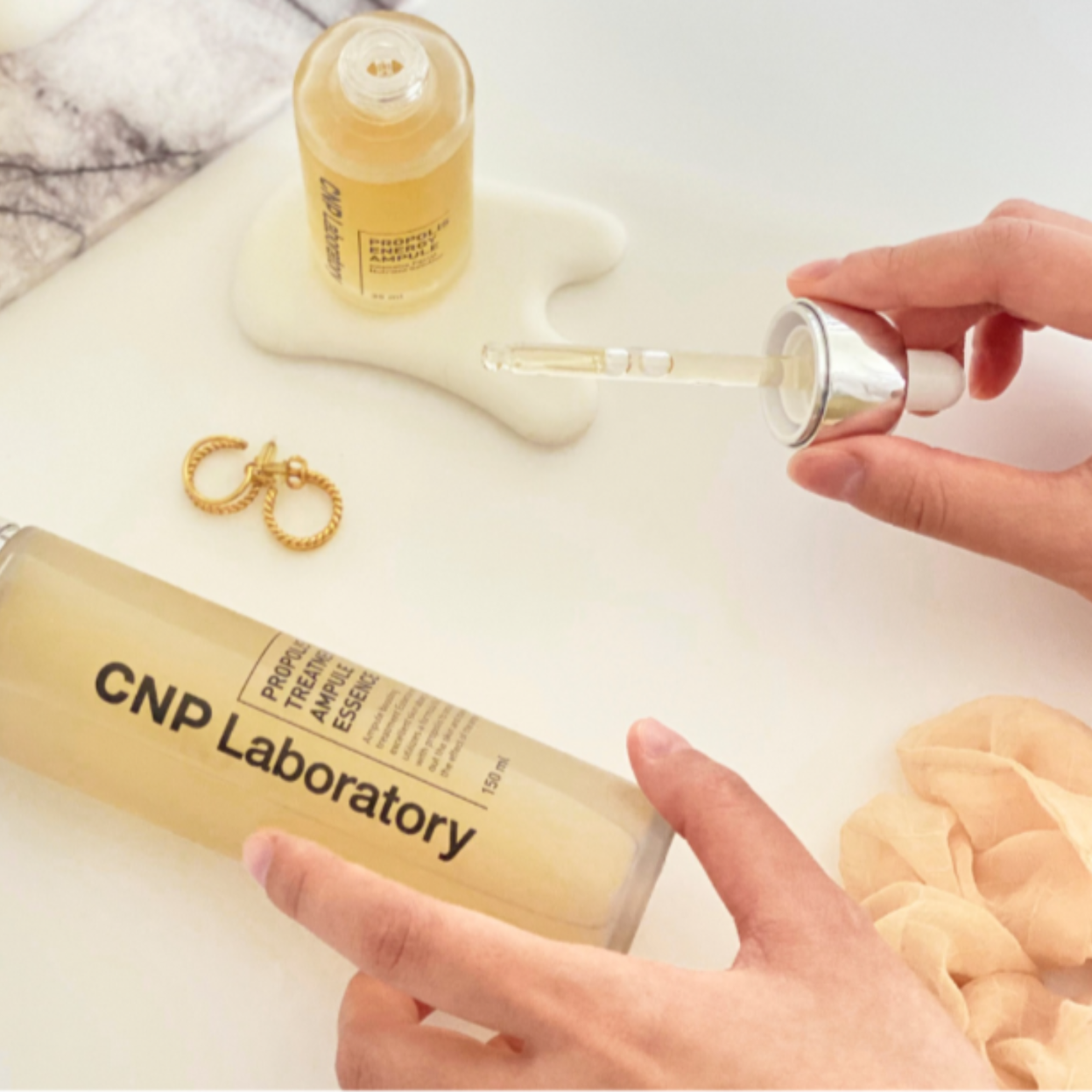 Left side: Hand holding a dropper with a bottle labeled CNP Laboratory Propolis Treatment Ampoule Essence, scattered beige shells, and a pair of gold hoop earrings. Right side: Various skincare products from CNP Laboratory, including a treatment ampoule, cream, and other bottles, on a white surface with a light-colored background.
