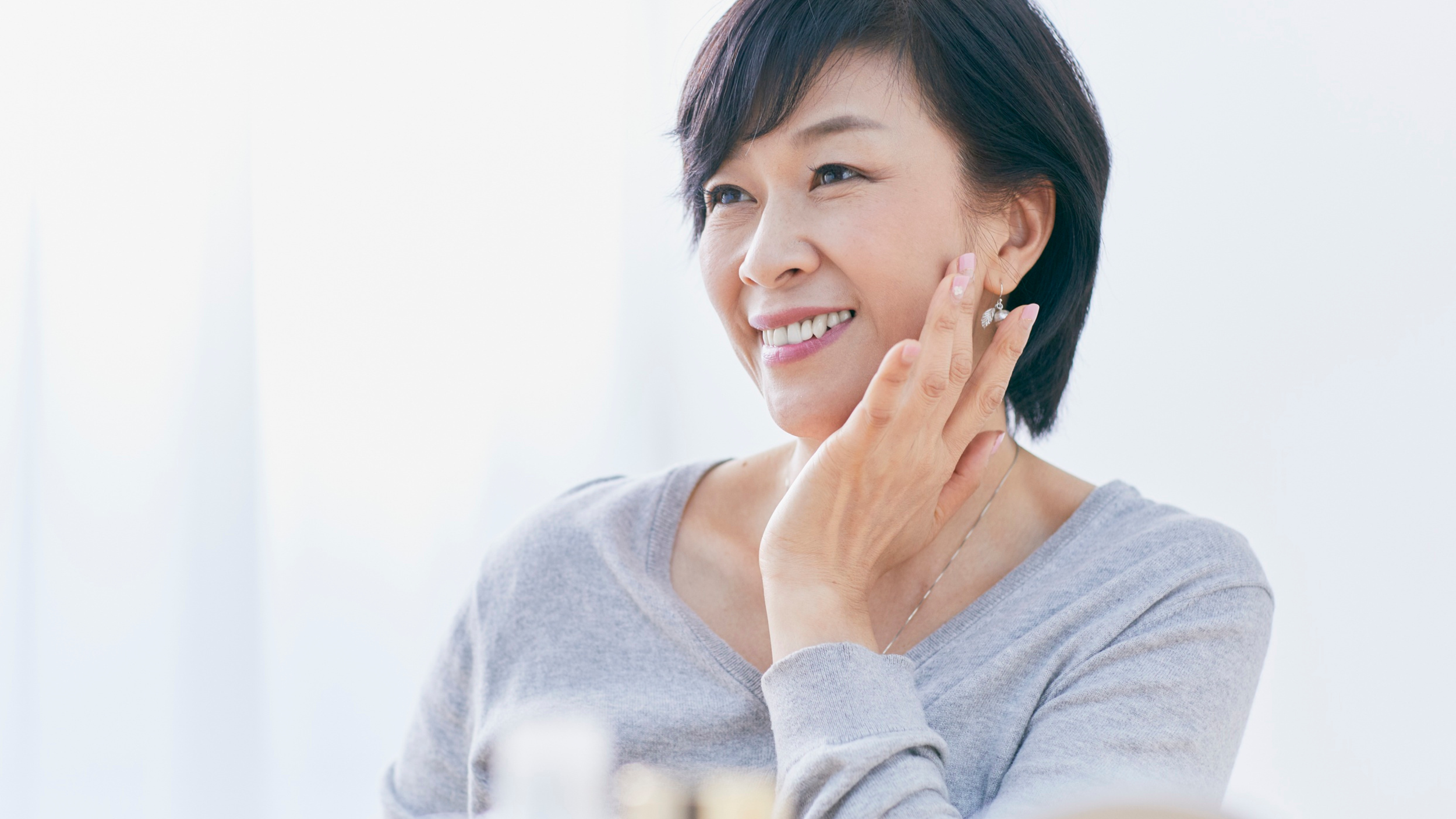 A woman smiling and touching her face, wearing earrings and a gray top, in a bright room.