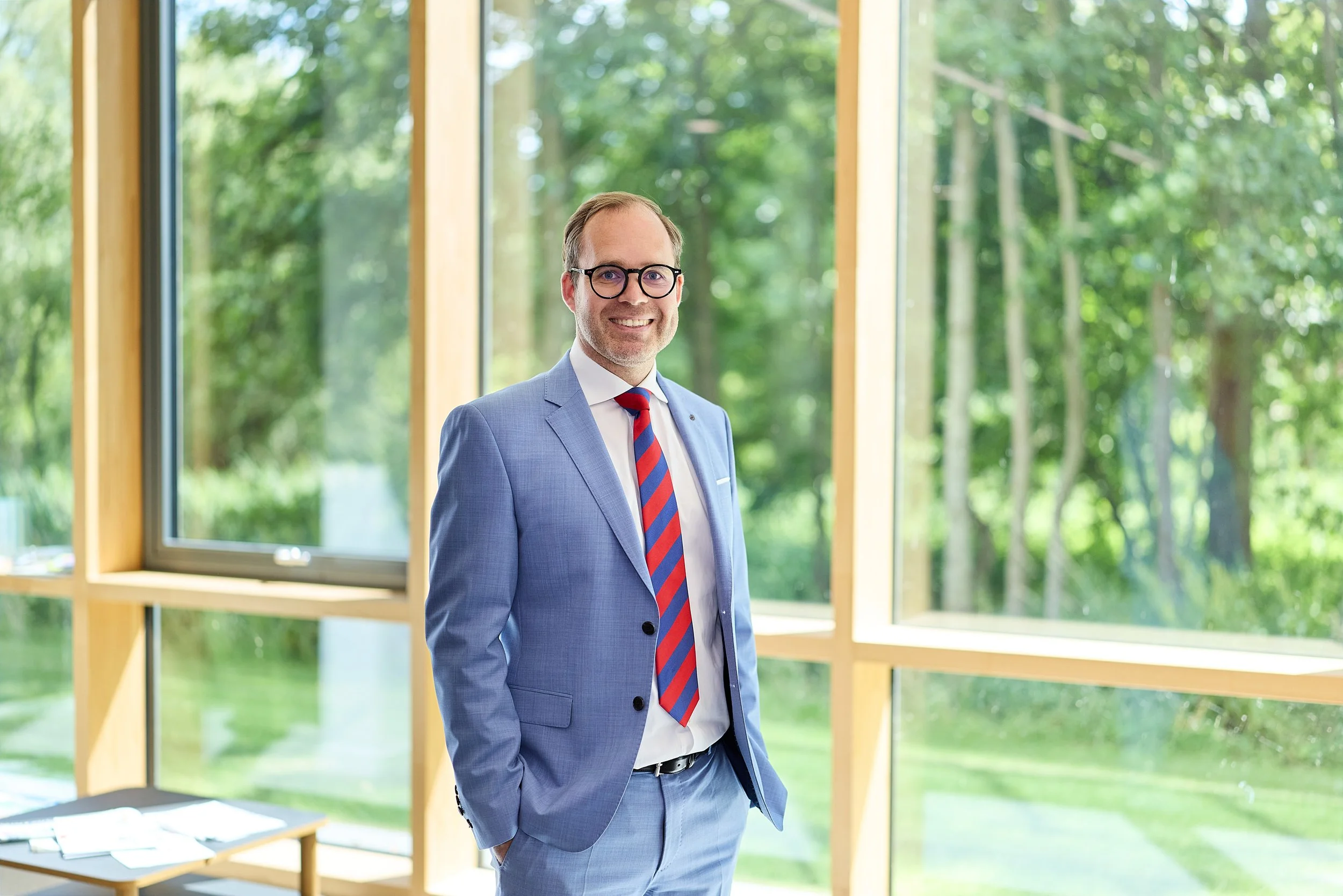 Ein Mann im blauen Anzug und roter, gestreifter Krawatte steht in einem Büro mit großen Fenstern und Blick ins Grüne, lächelnd in die Kamera.