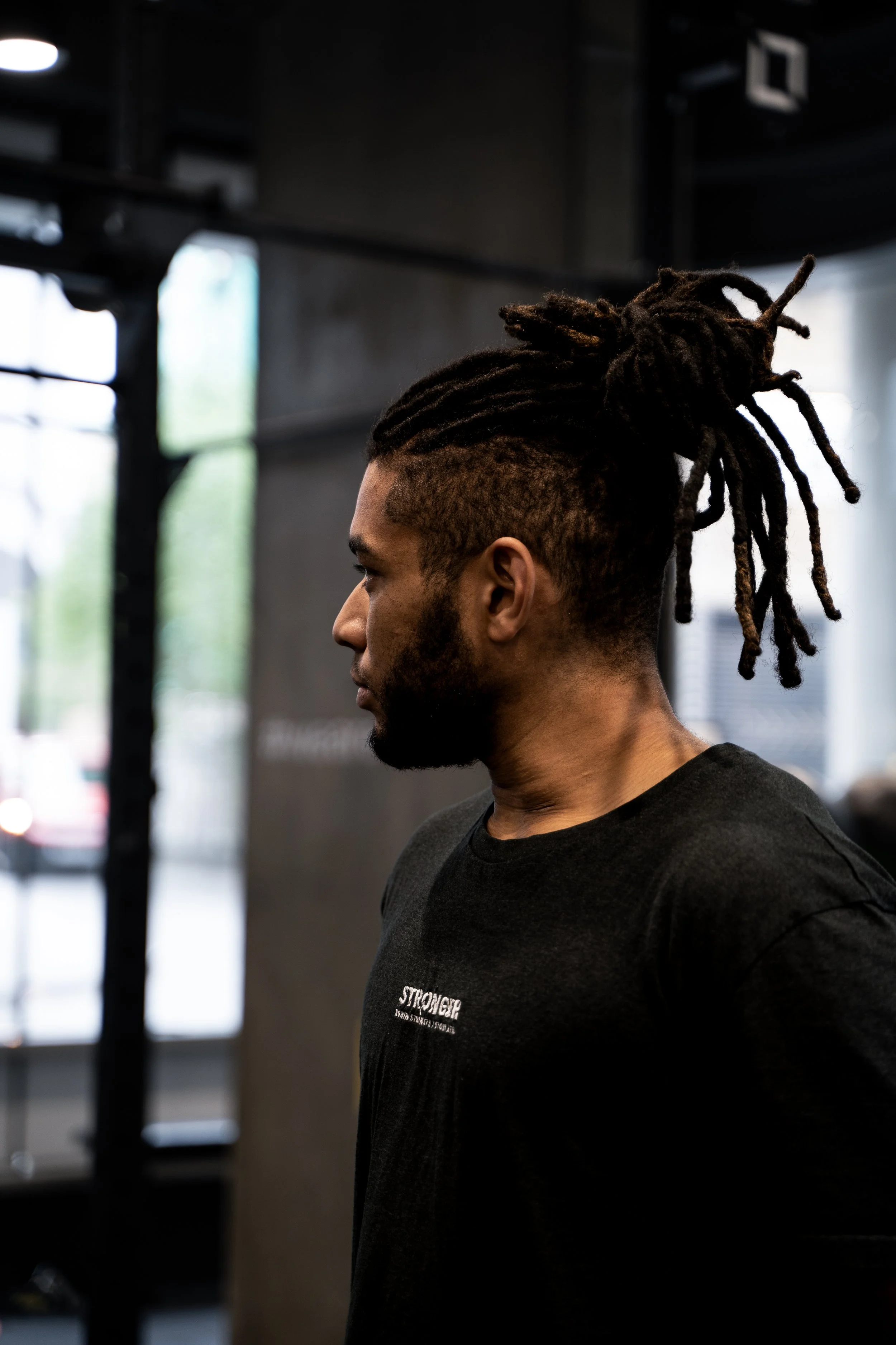 A man with dreadlocks tied up in a bun, wearing a black shirt, standing indoors in profile view.