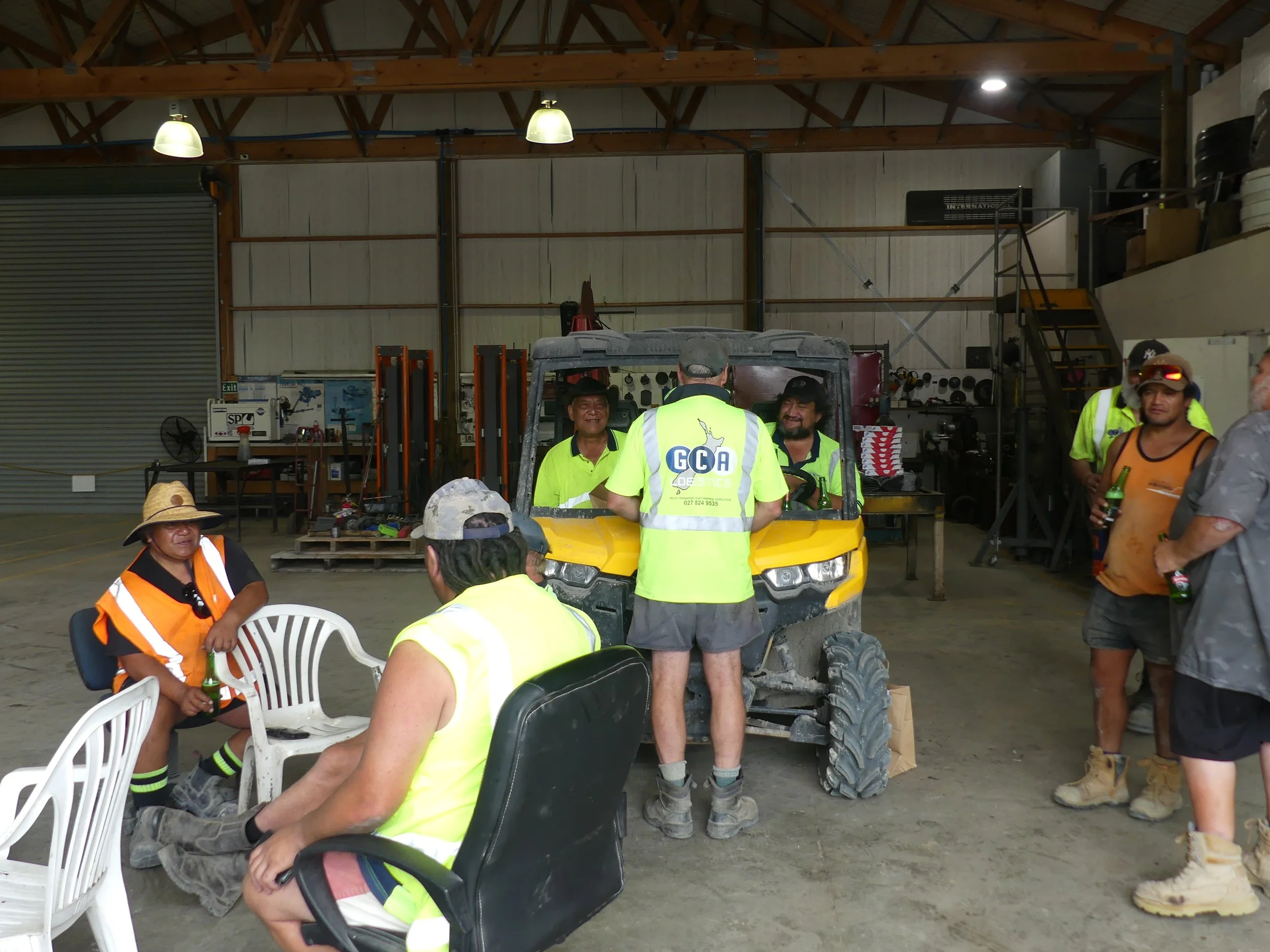 Group of people, some in reflective vests, inside the GCA Logistics yard with a yellow utility vehicle in the center, some sitting and some standing, engaged in conversation.
