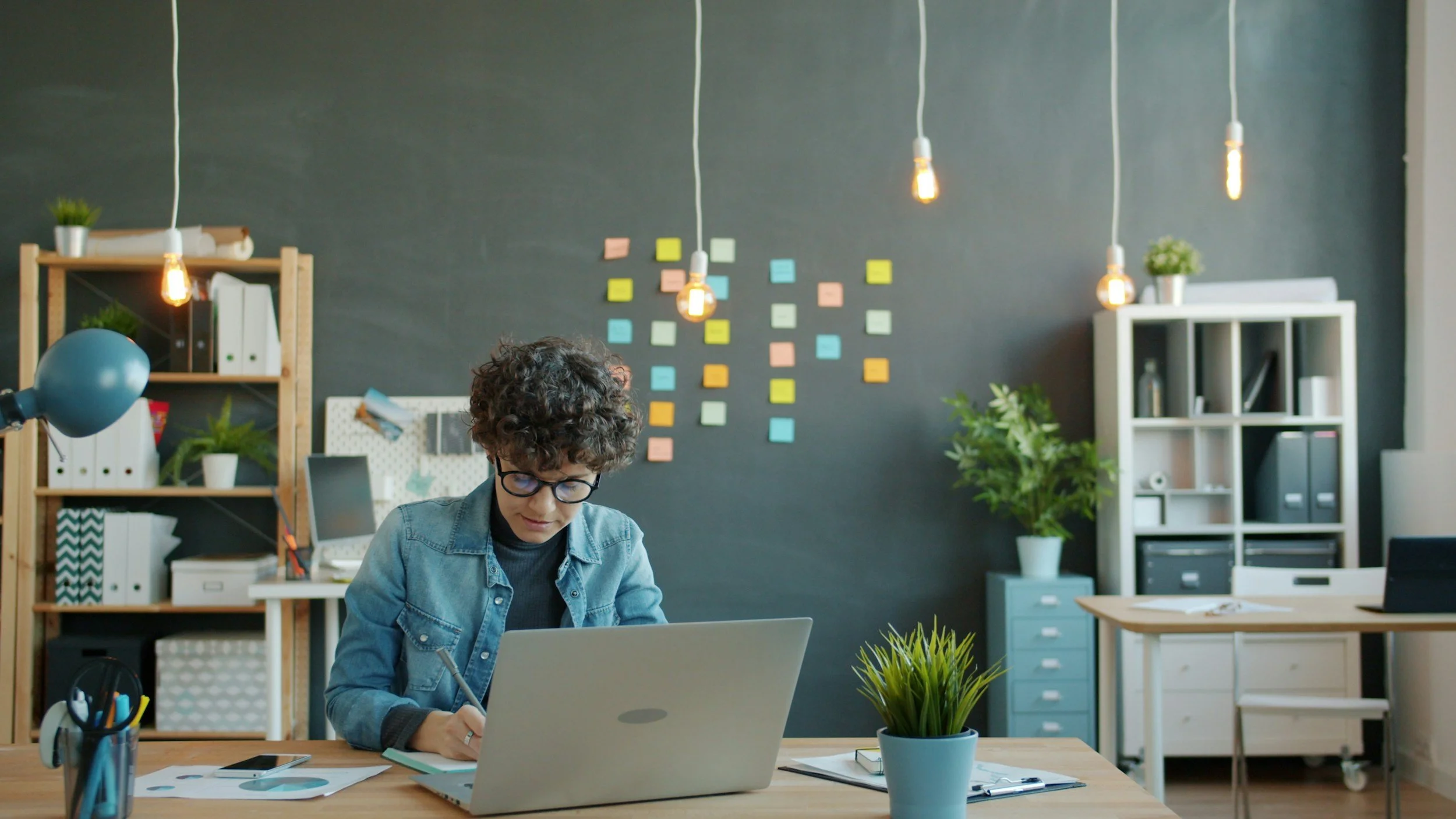 Person with short curly hair and chambray shirt sitting at a desk with a laptop and plant. The dark wall behind has brightly-colored post-it notes with ideas for a personal statement story.