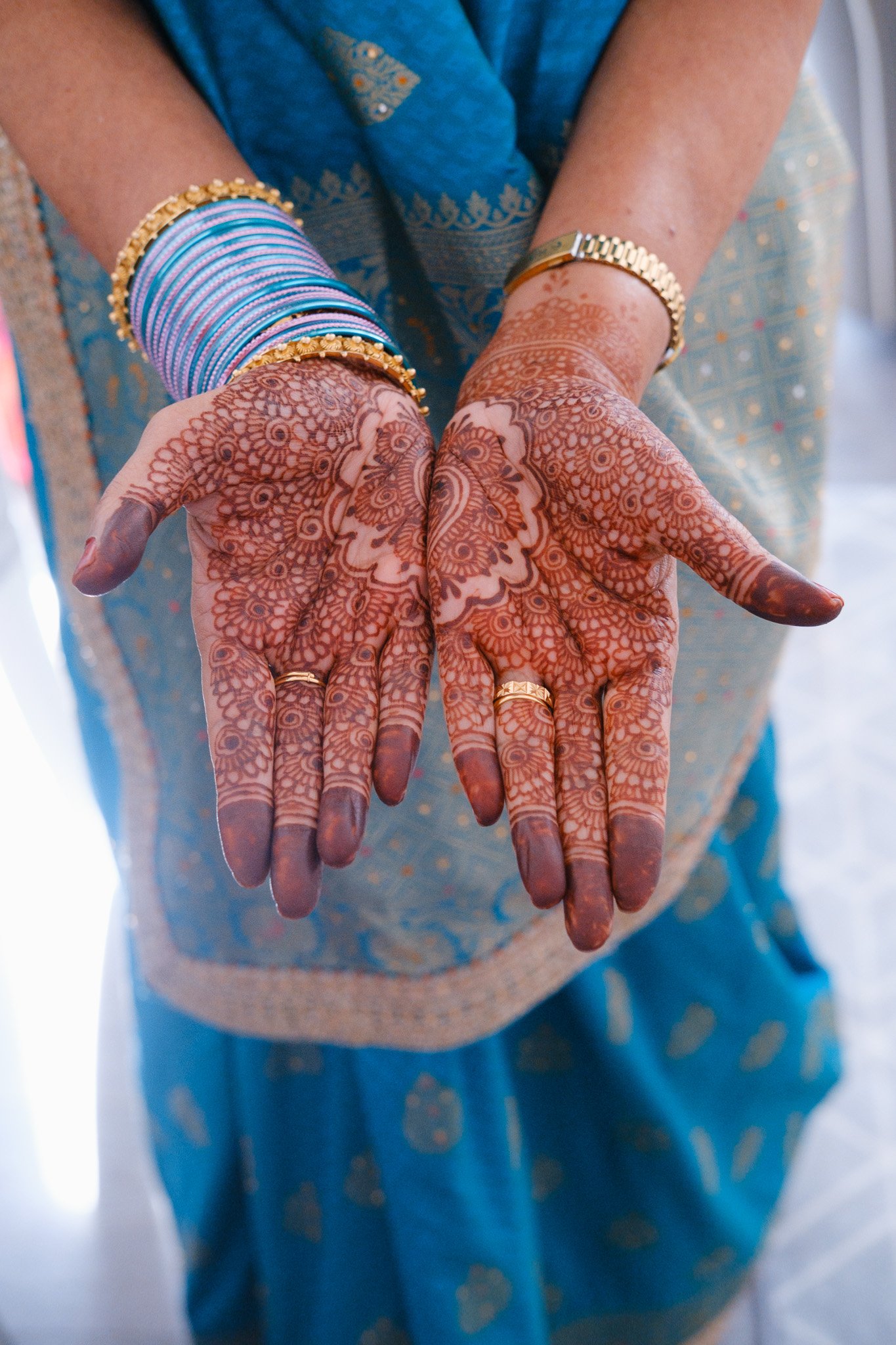 Close-up of hands with intricate henna designs, wearing blue and gold bangles, against a background of blue and gold fabric.