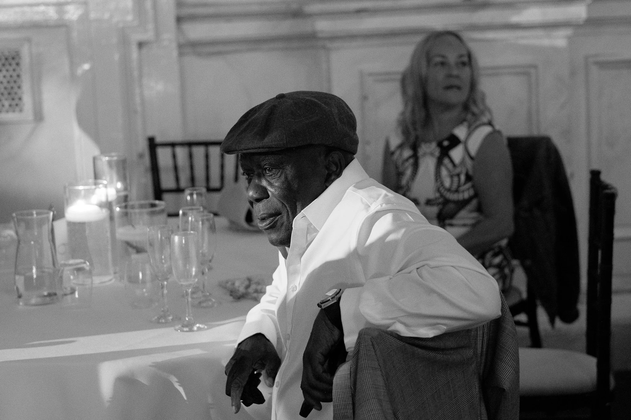 Black and white image of an older man wearing a flat cap and white shirt, sitting at a table with empty glassware, with a woman in the background.