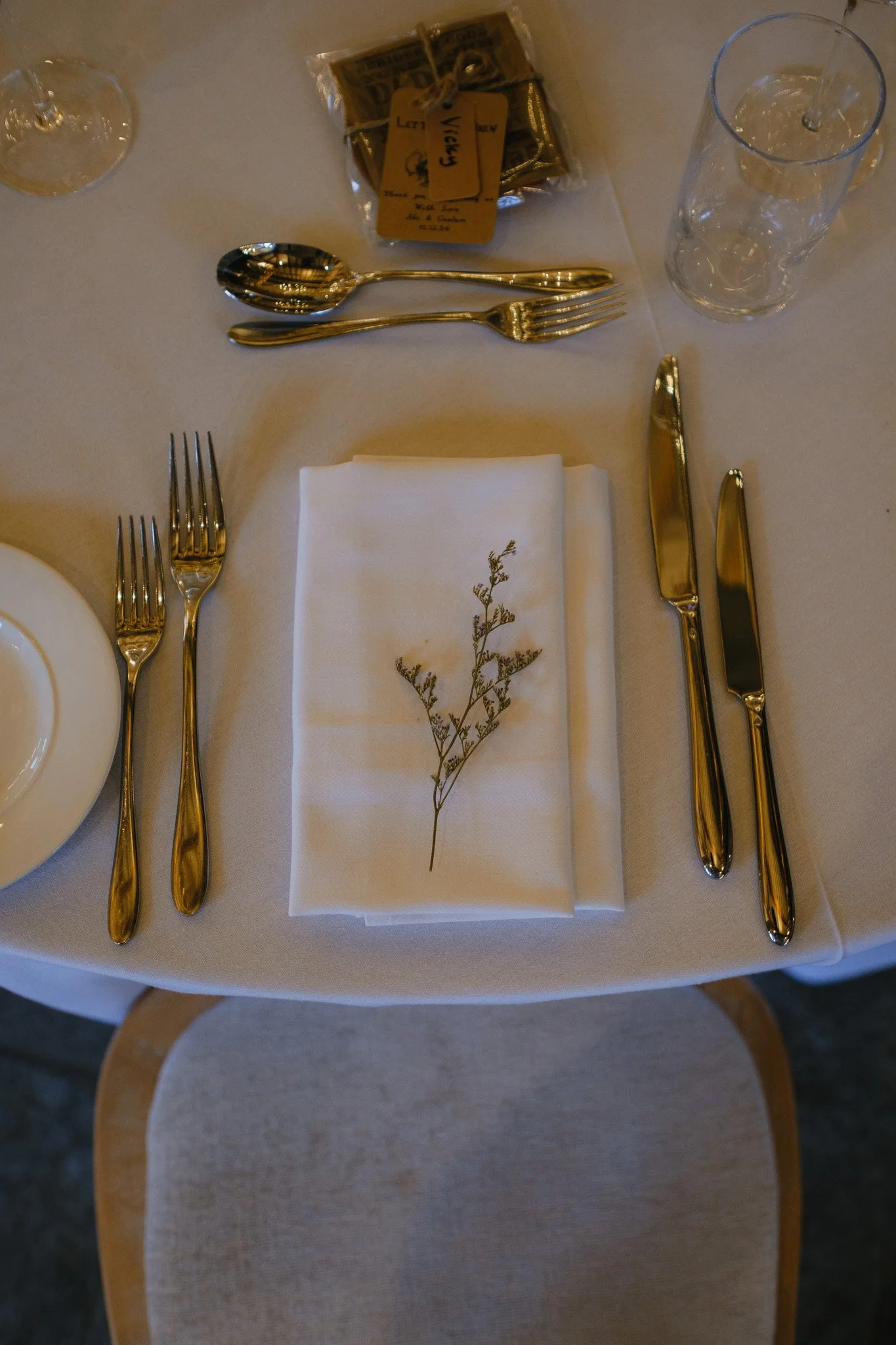 Elegant table setting with silverware, white napkins, a sprig of dried flowers, a glass, and a small wrapped gift on a white tablecloth.