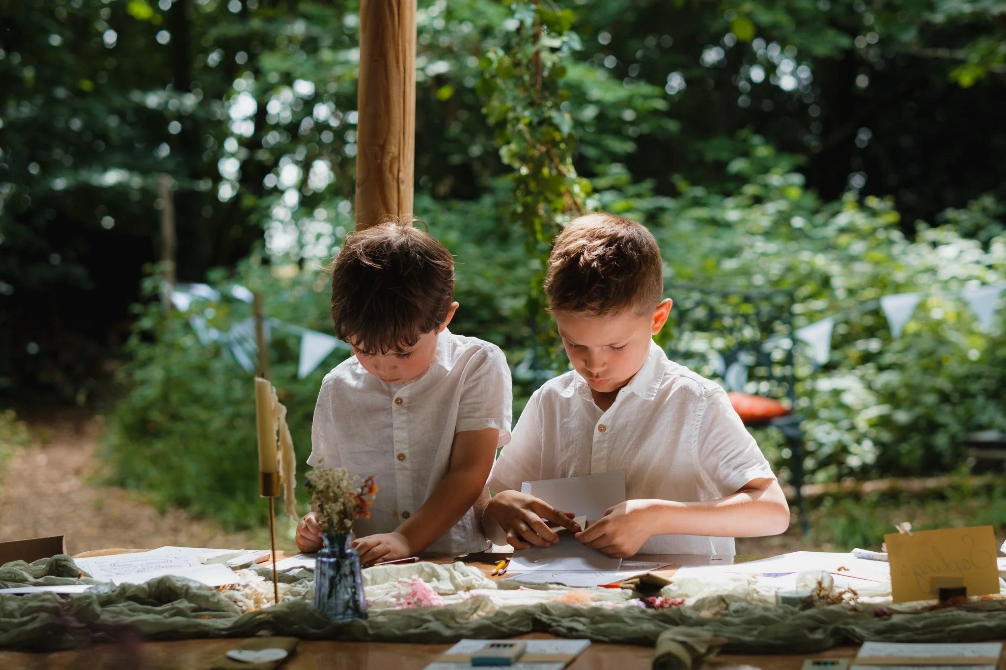 Two young boys in white shirts are engaged in activities at an outdoor table decorated with flowers and items, in a forested area with trees and greenery in the background.