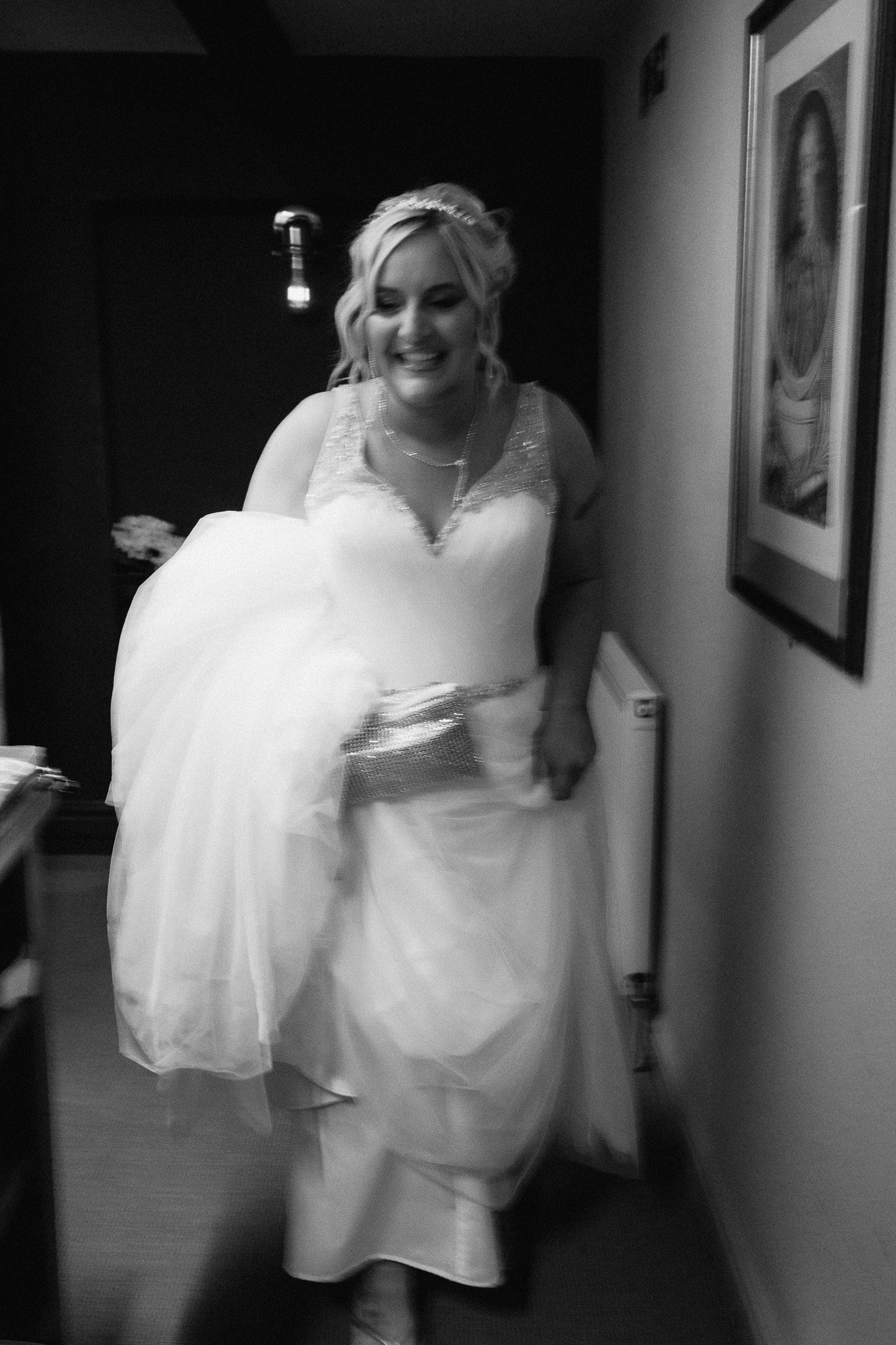 Blurred black and white photo of a bride in a wedding dress smiling and holding her gown in a hallway with framed artwork on the wall.