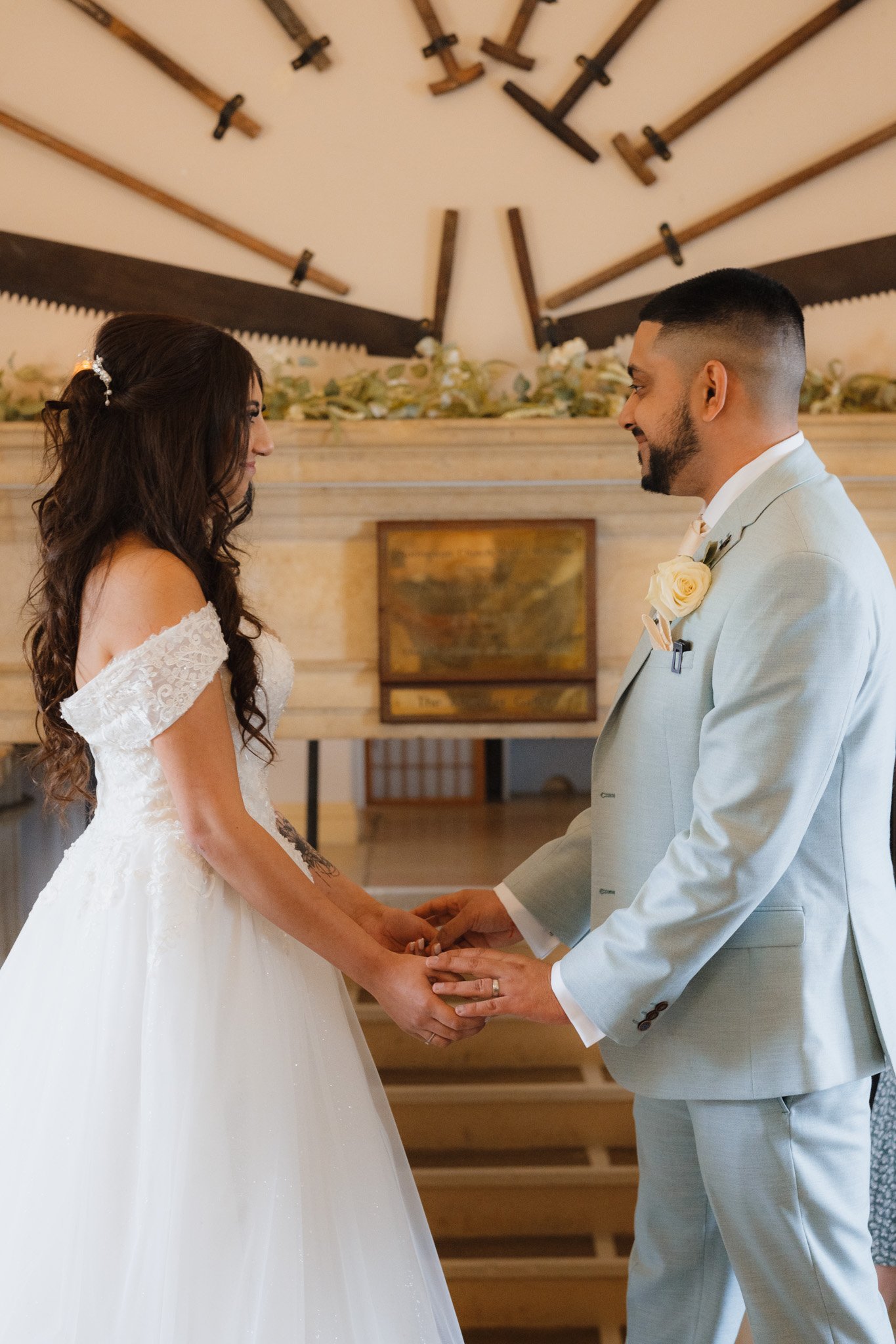 Bride and groom holding hands during a wedding ceremony, with a decorative wall of vintage tools in the background.