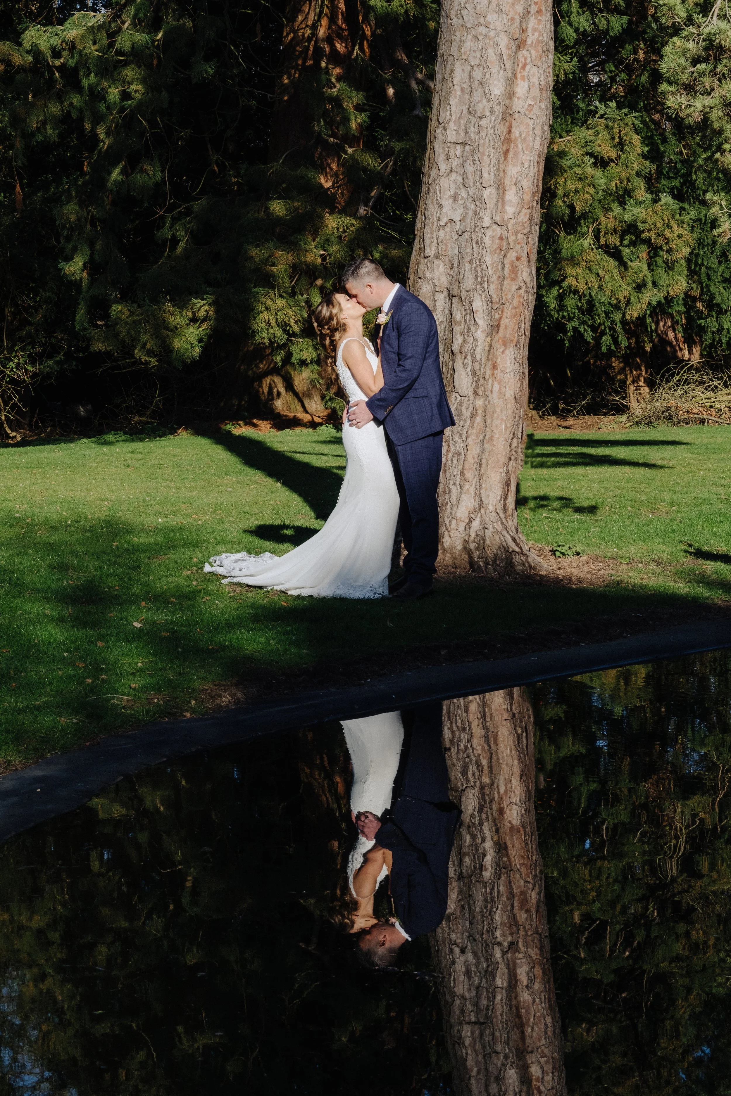 A bride and groom kissing by a tree with a reflection in water.