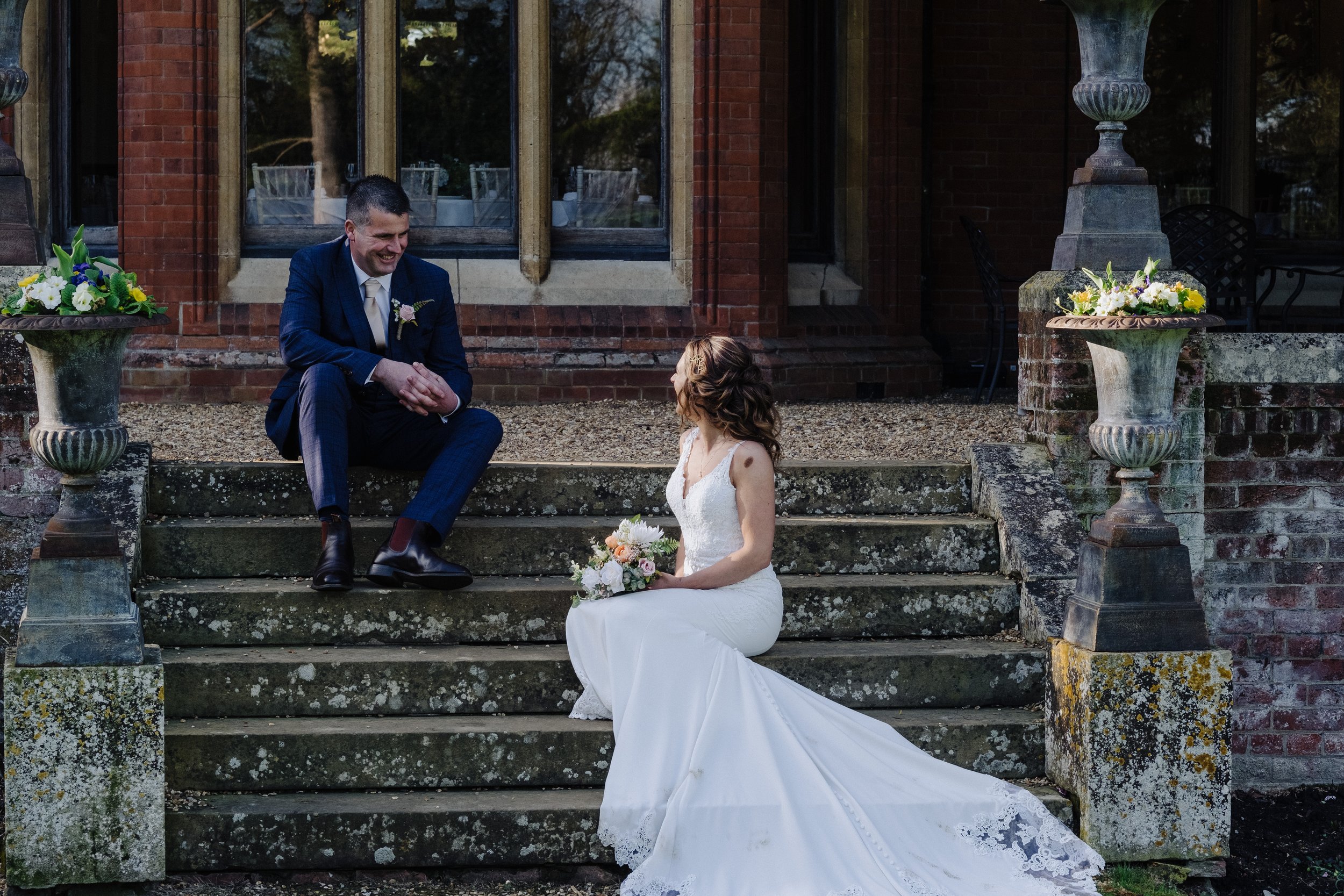 A bride and groom sit on stone steps outside a building. The groom is wearing a blue suit and the bride is in a white wedding dress, holding a bouquet. There are two large flower-filled urns on either side of the steps.