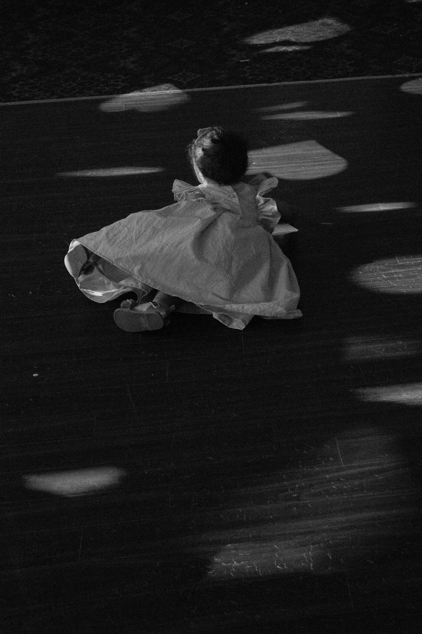 Black and white image of a child in a dress sitting on a wooden floor with light patterns.
