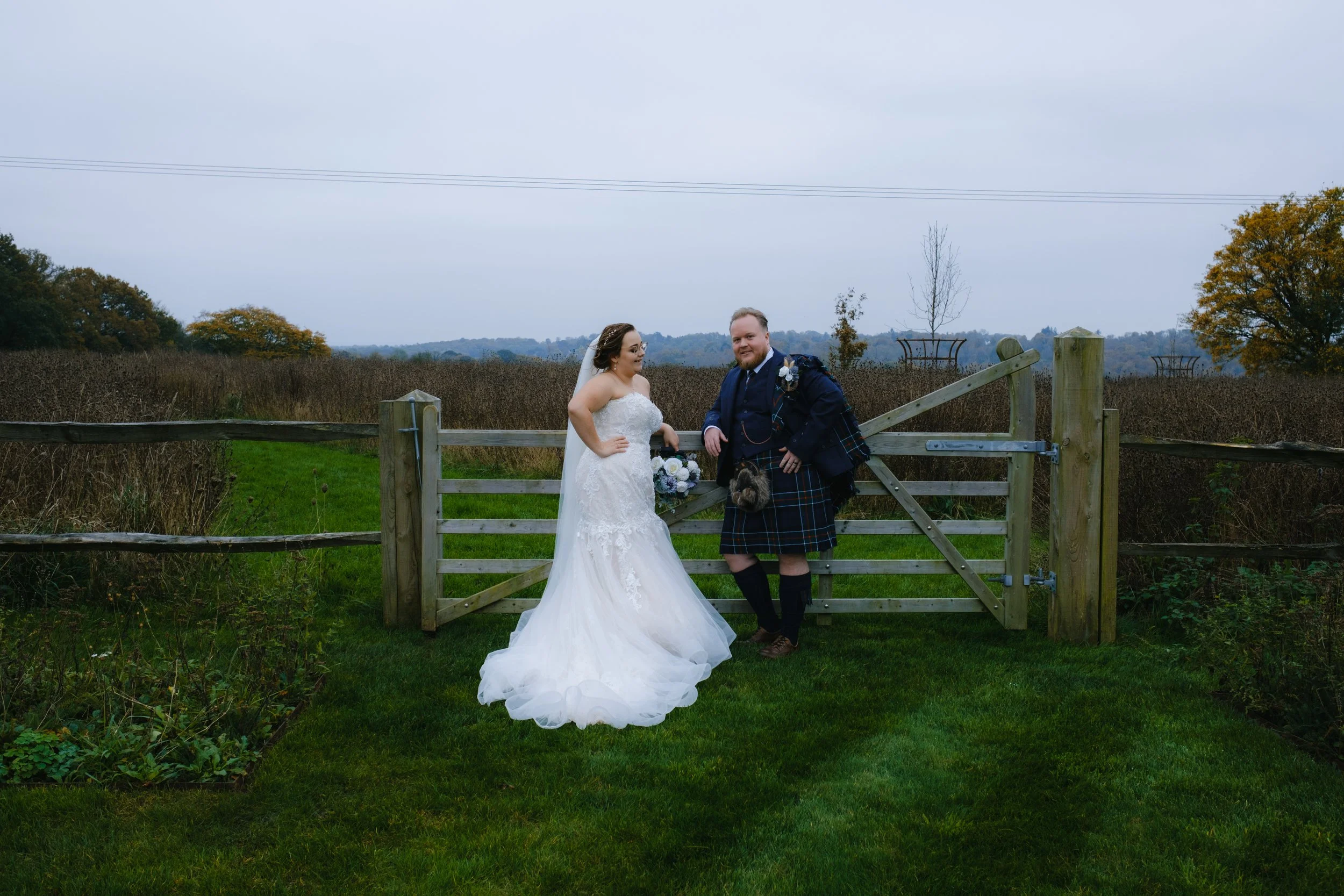 Bride in white gown and groom in Scottish kilt standing by a wooden gate in a grassy field.