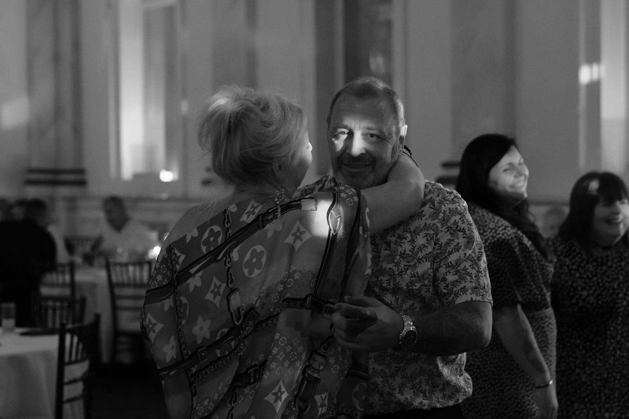 A black and white photo of people dancing at a social event. A man and woman are in the foreground, with the woman’s arm around the man's neck, smiling. Other attendees are seen in the background.