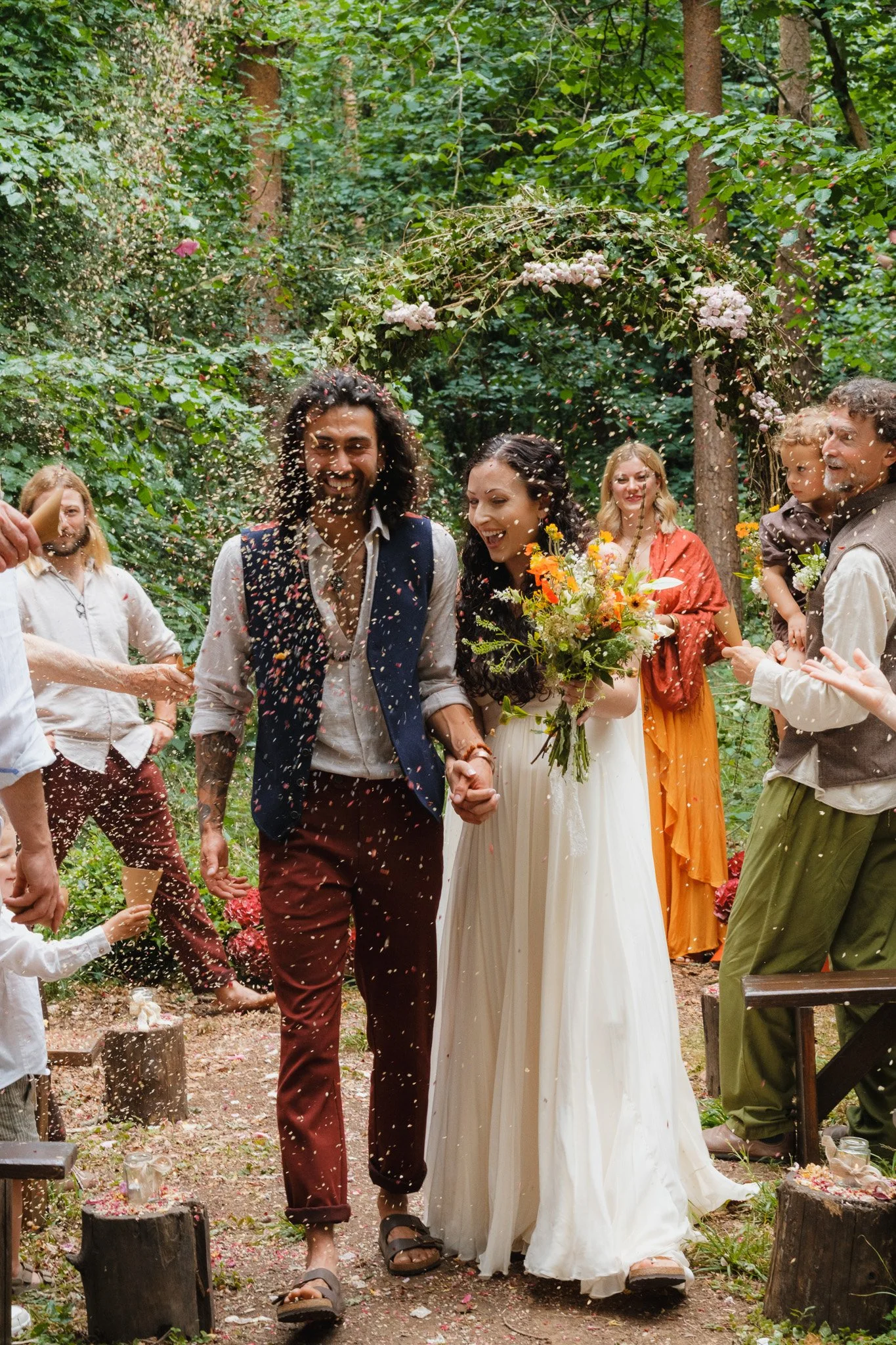 A newlywed couple holding hands, smiling, and walking through a forested outdoor wedding ceremony surrounded by friends and family, with confetti being thrown in celebration.