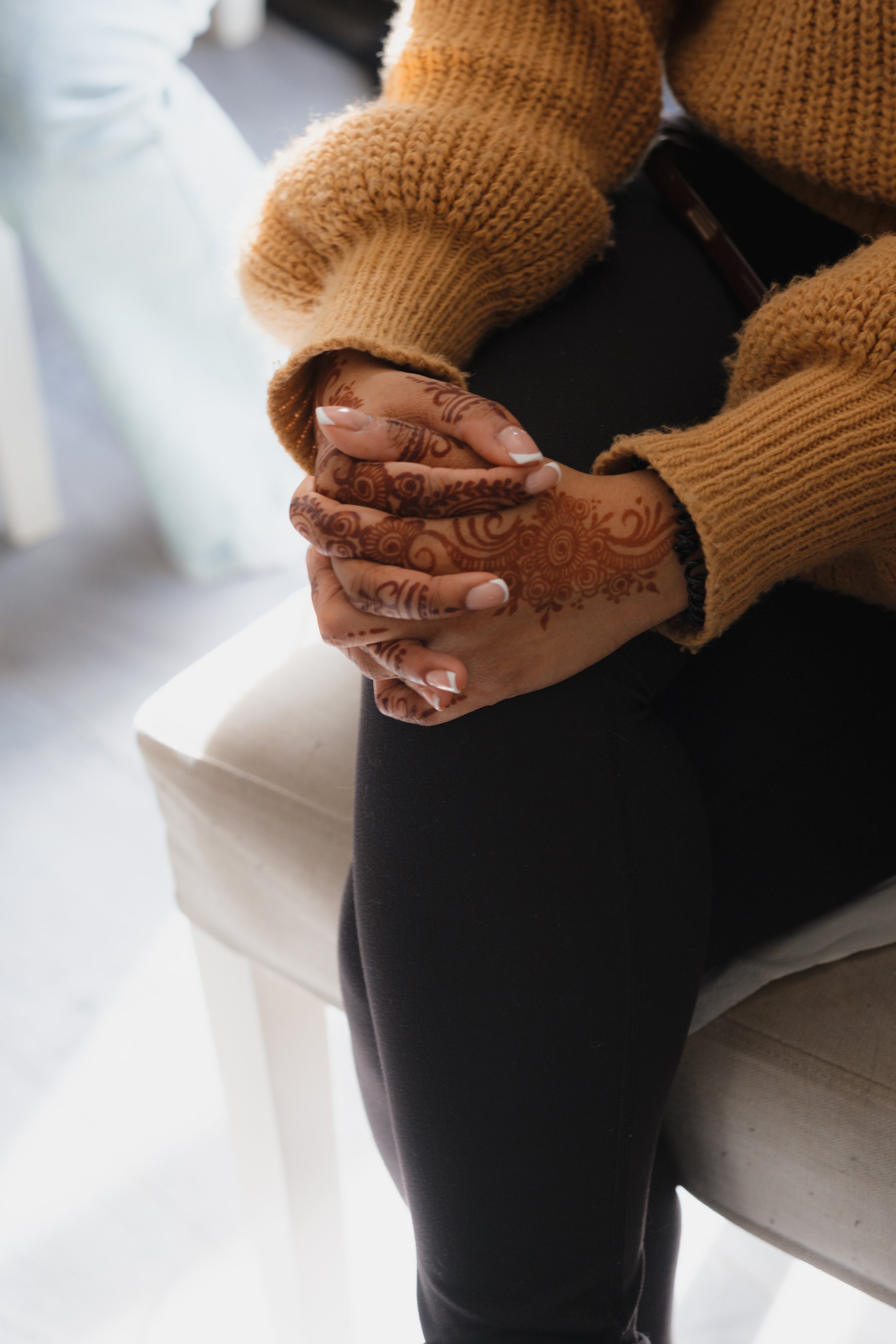 Close-up of folded hands with henna designs on seated person wearing a brown sweater and black pants.