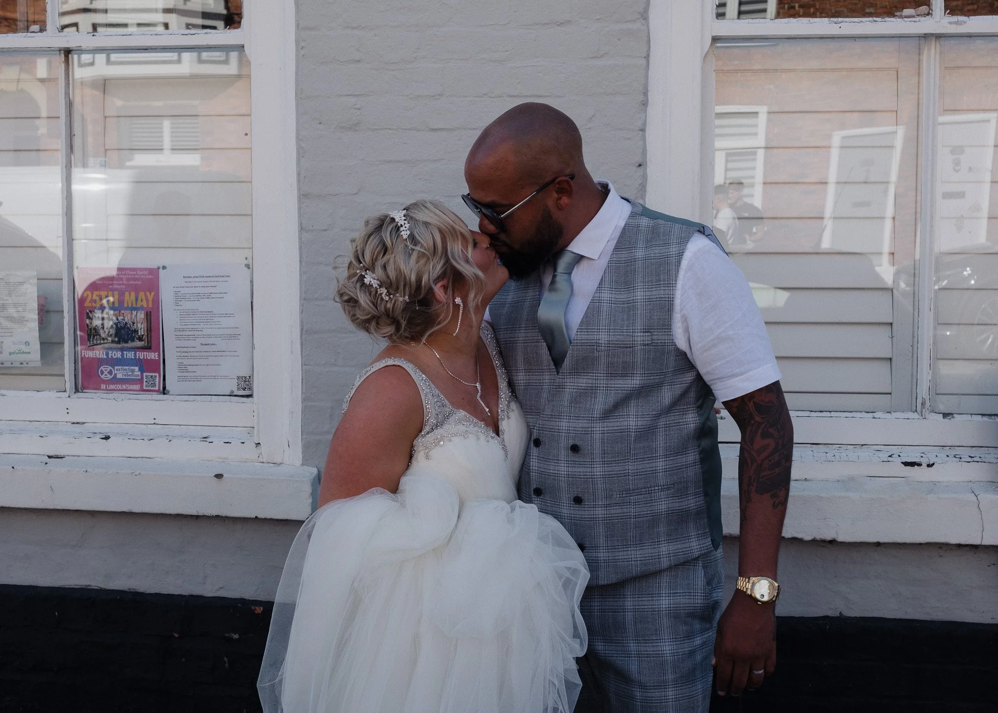 A bride and groom kissing in front of a white brick wall, with reflections visible in the window behind them. The bride is in a white wedding dress, and the groom is wearing a gray plaid vest.