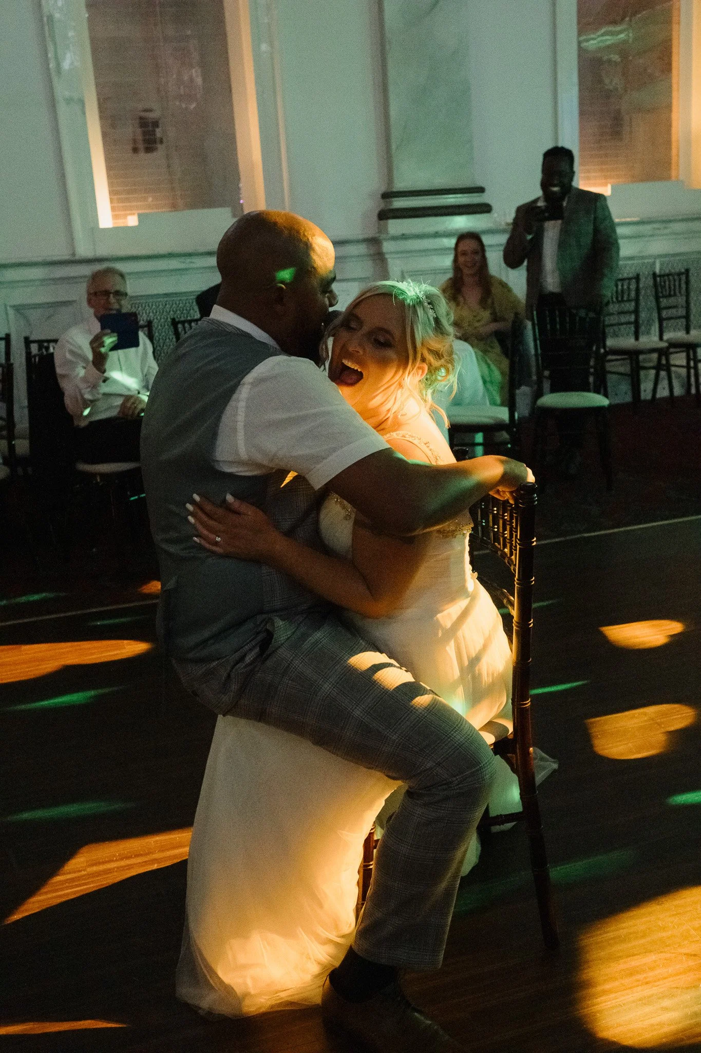 Couple dancing intimately at a wedding reception, with guests seated and watching in a dimly lit room with green lighting.