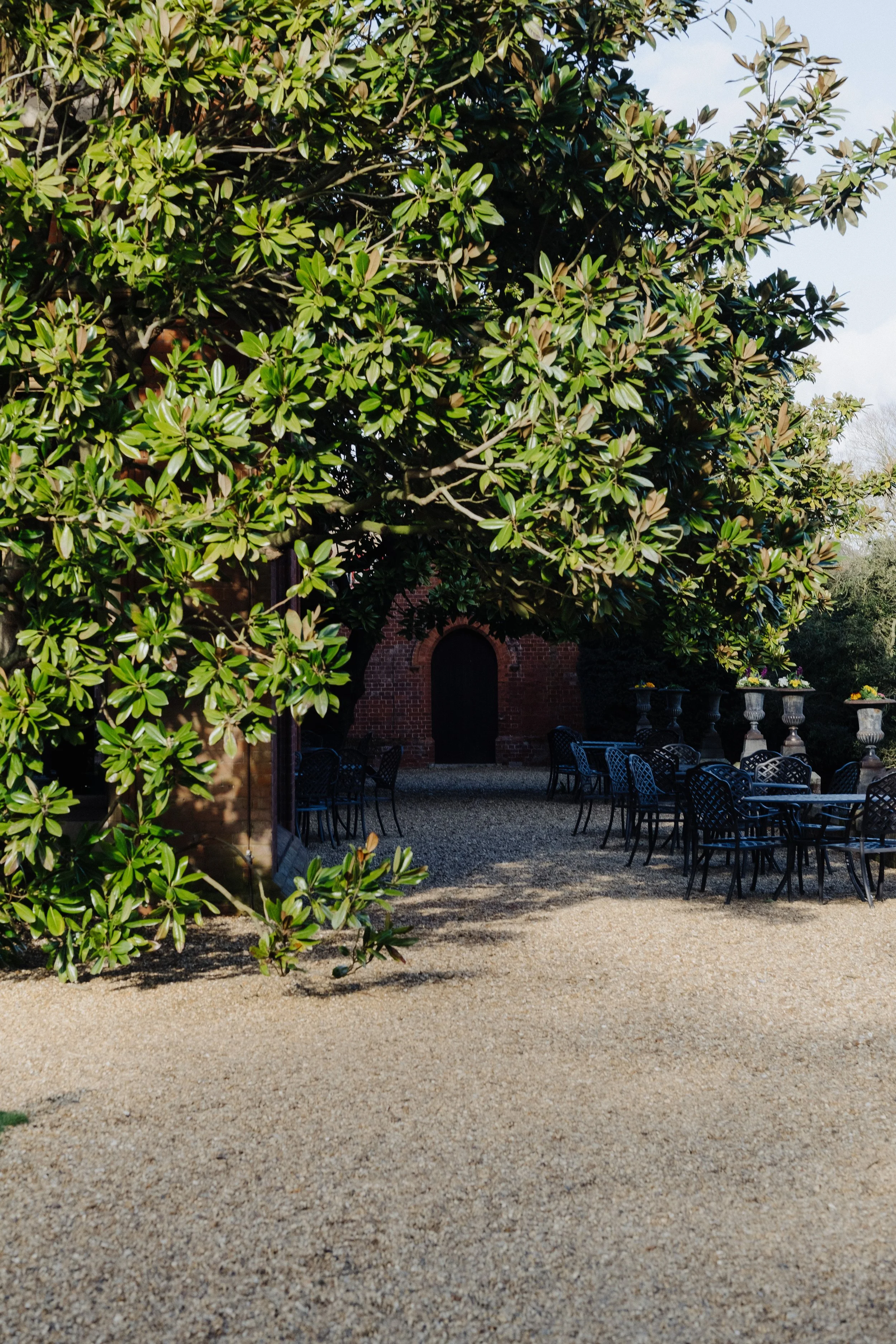Outdoor patio with metal tables and chairs, shaded by a large tree with dense green foliage, a brick wall with an arched door in the background.