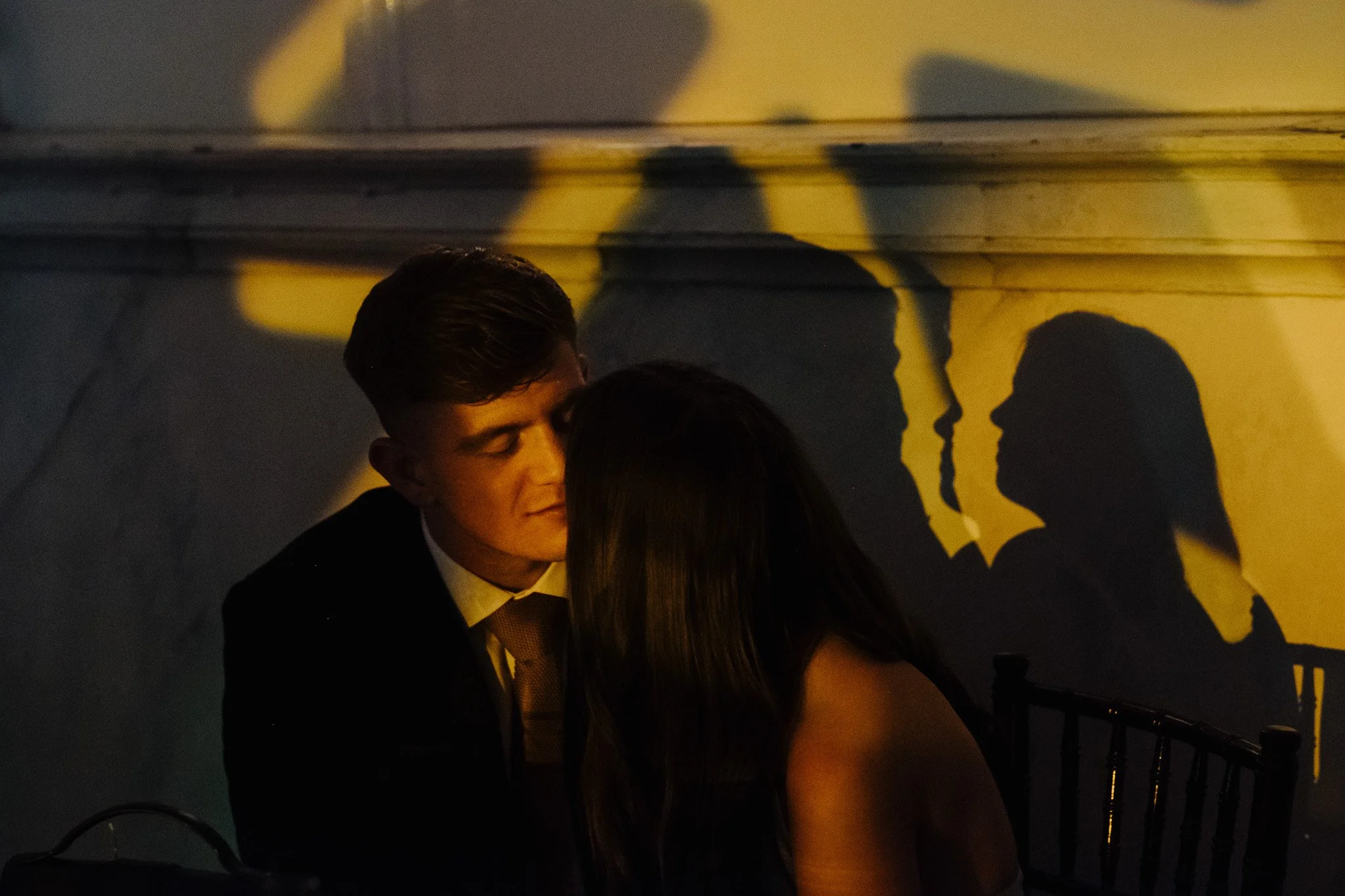 A couple sitting closely with shadows on a wall behind them, creating silhouettes in a dimly lit setting.