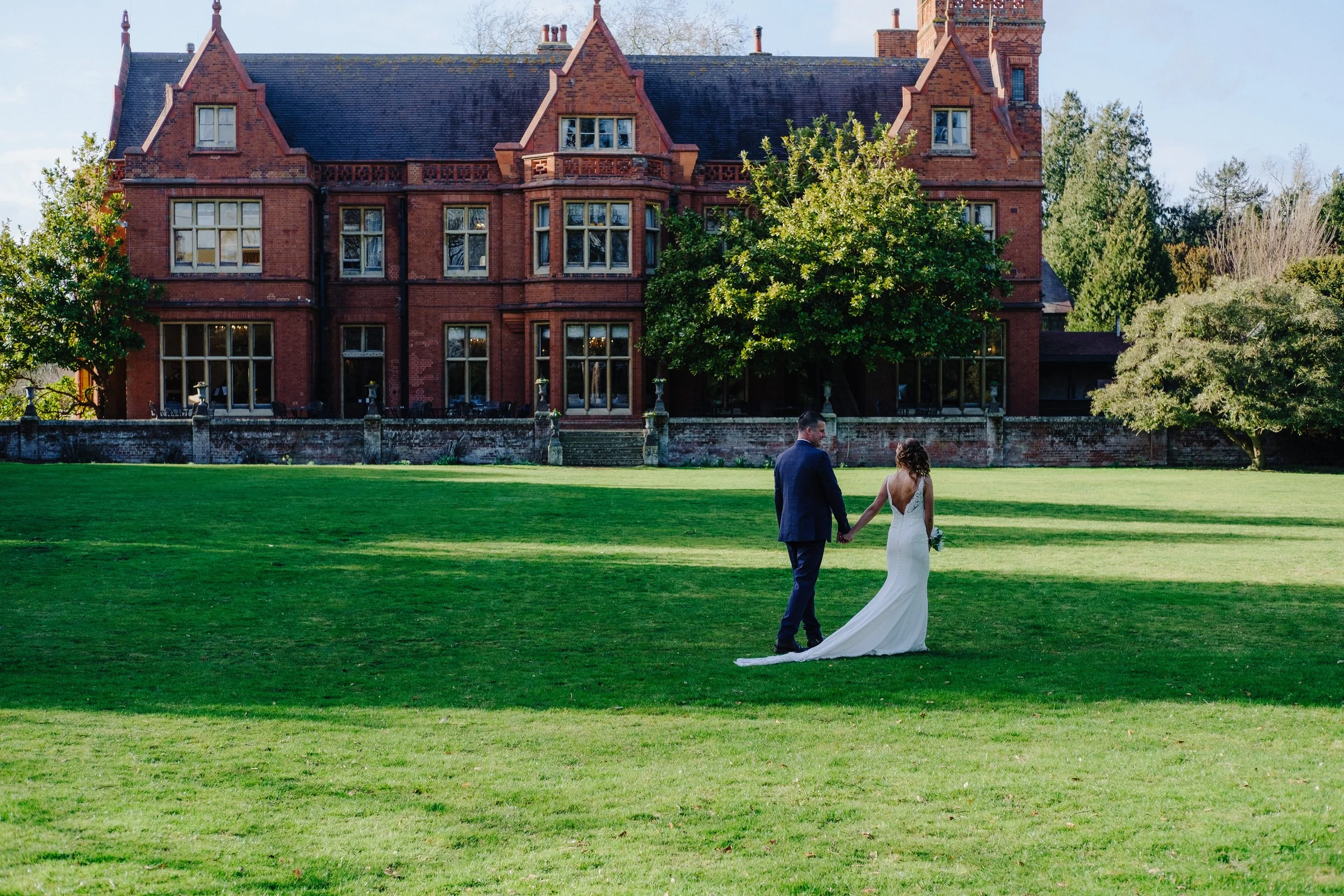 A couple walking on a grassy lawn towards a large, old brick mansion with trees around it.
