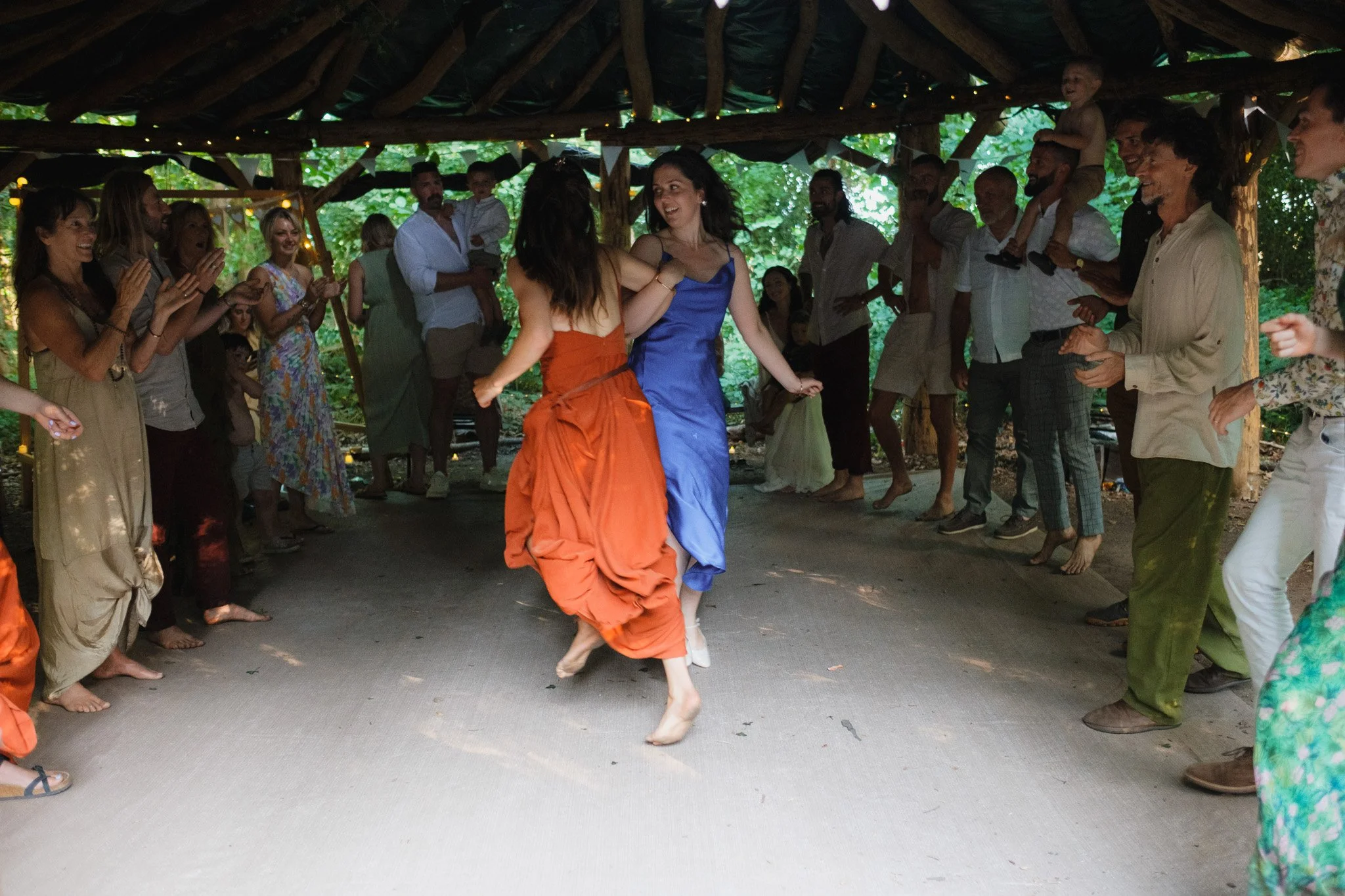 People dancing in a circle at a celebration or party in an outdoor wooden rustic shelter surrounded by greenery.