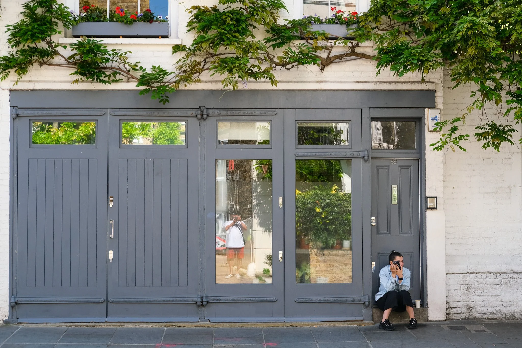 Person sitting outside a gray-painted urban house with vine-covered facade, door, and window. Reflection in glass shows photographer. Flower boxes above doors.