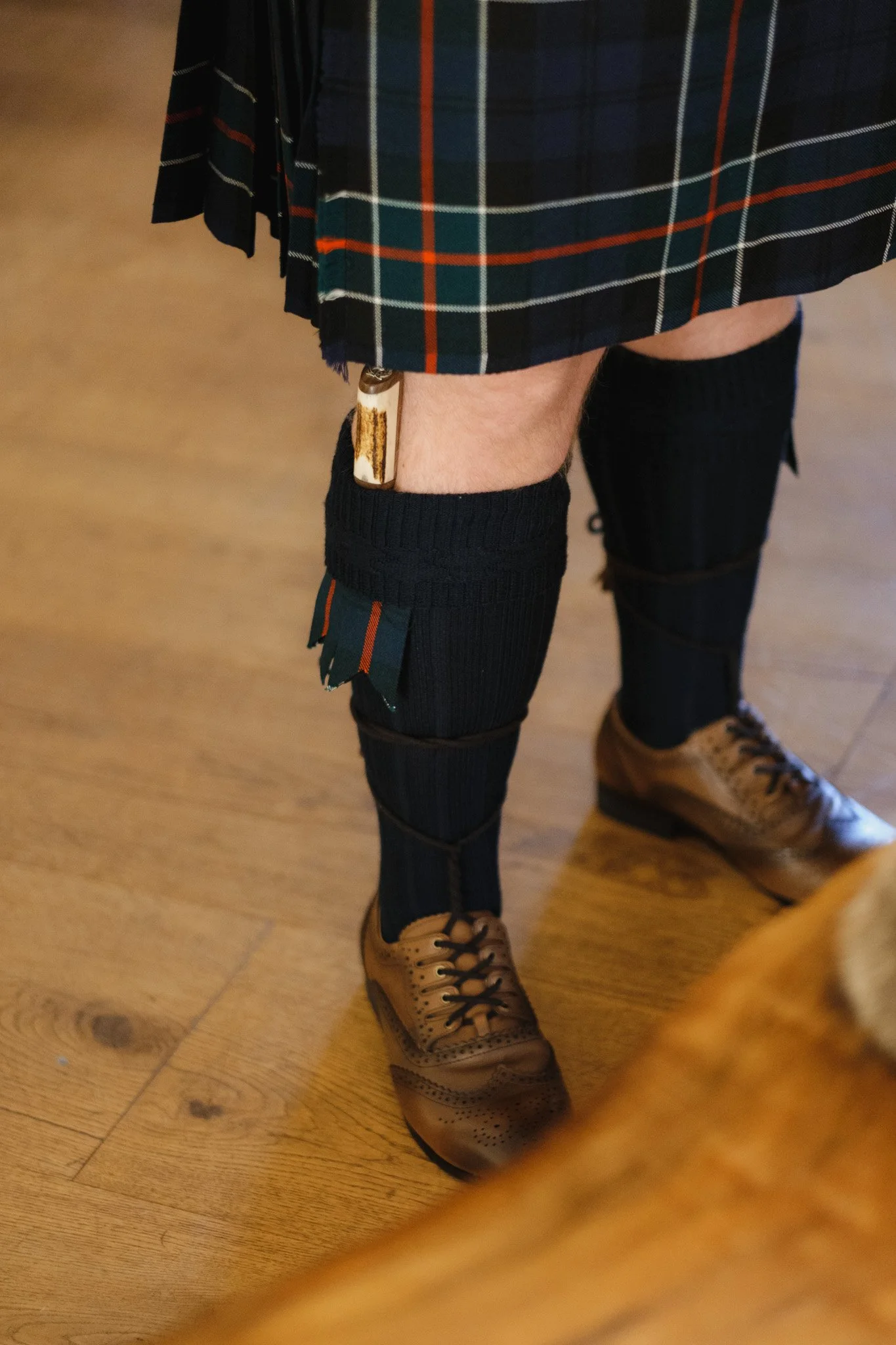 Person wearing a dark tartan kilt with matching socks and brown leather shoes on a wooden floor.