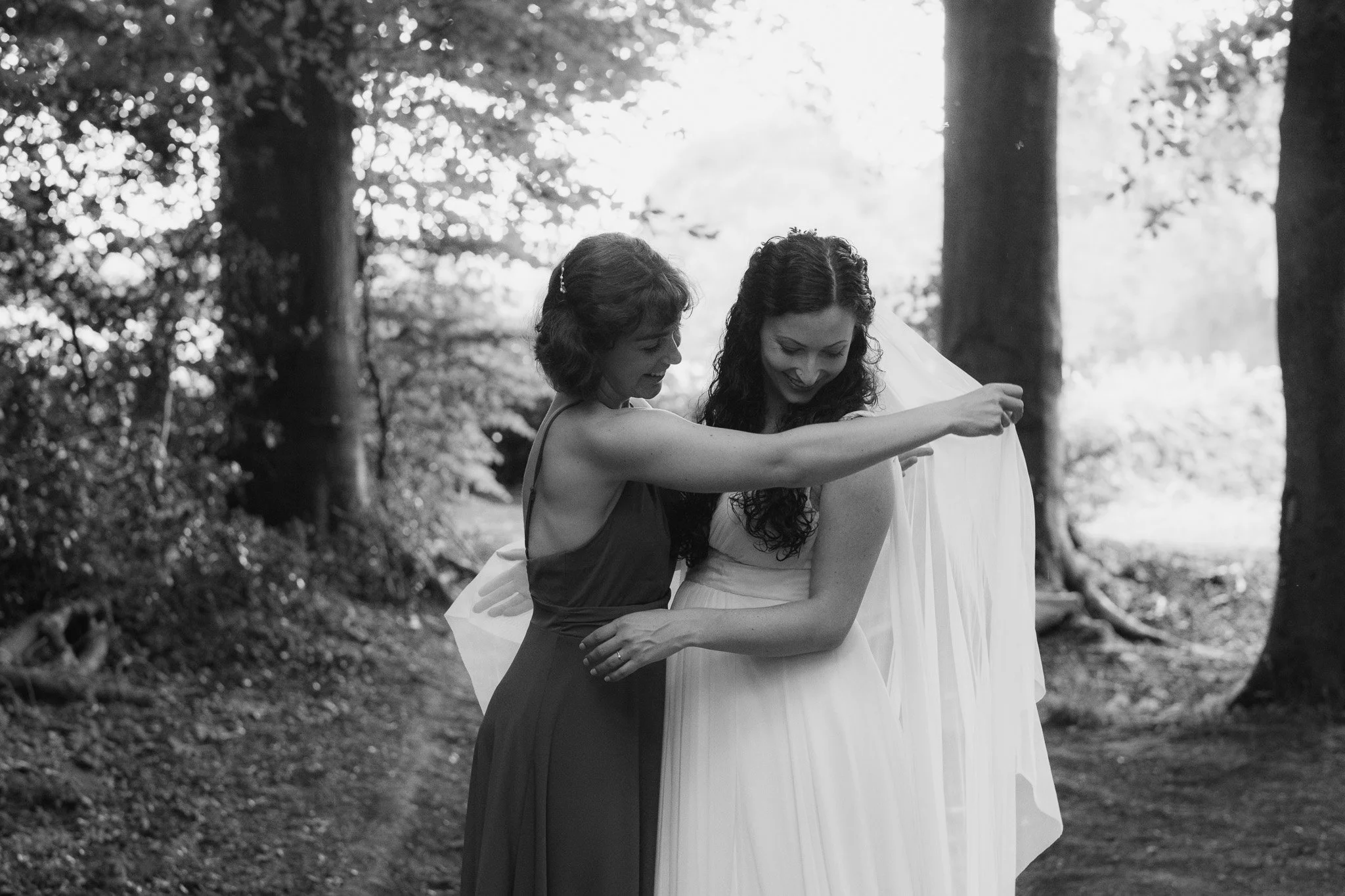 Two women, one in a dark dress and the other in a wedding dress, standing together outdoors in a forest, smiling and sharing a joyful moment.