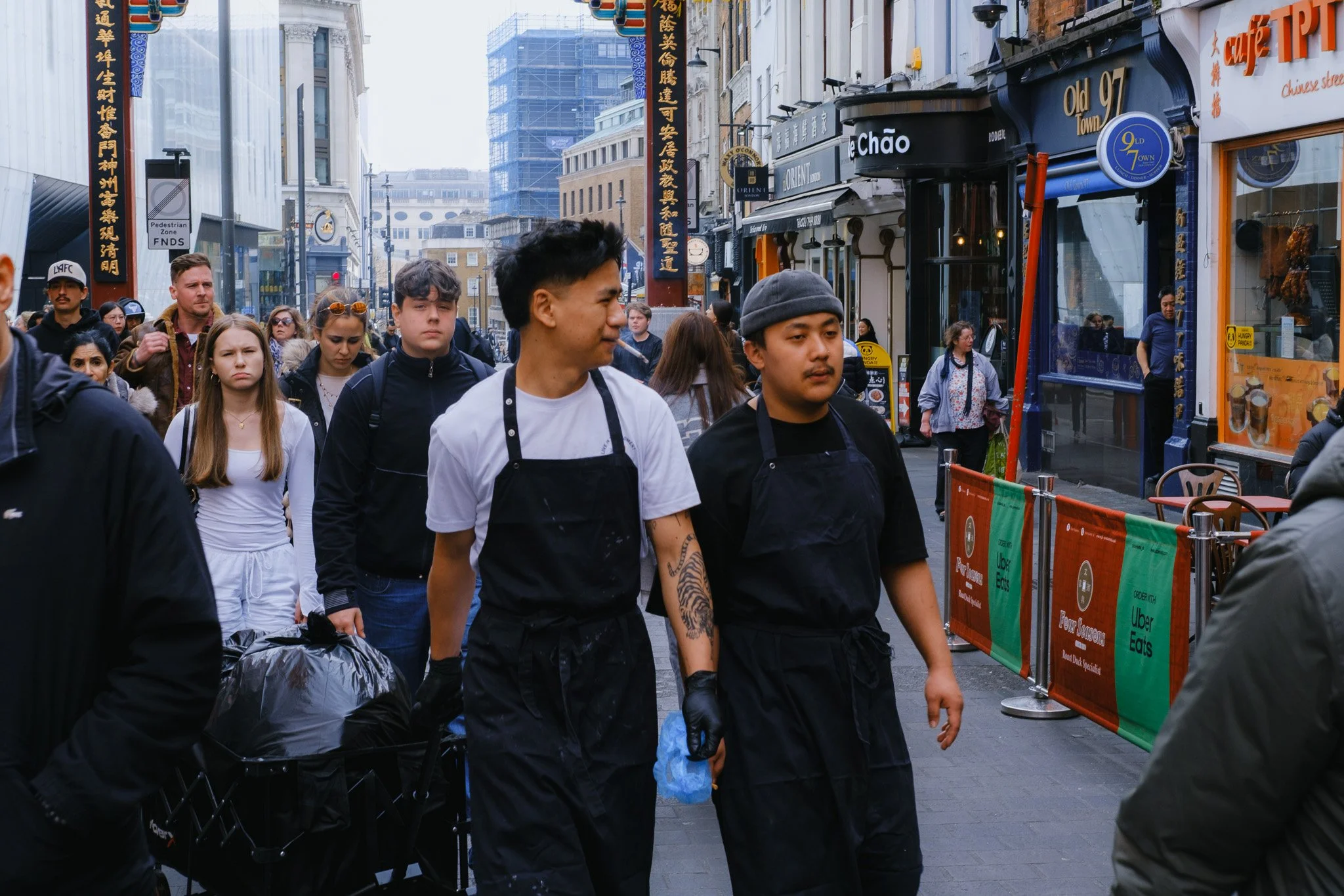 People walking on a busy street near restaurants and shops in a city, with some individuals wearing aprons and gloves; a decorative pillar with Chinese characters is visible, suggesting a location in a Chinatown area.