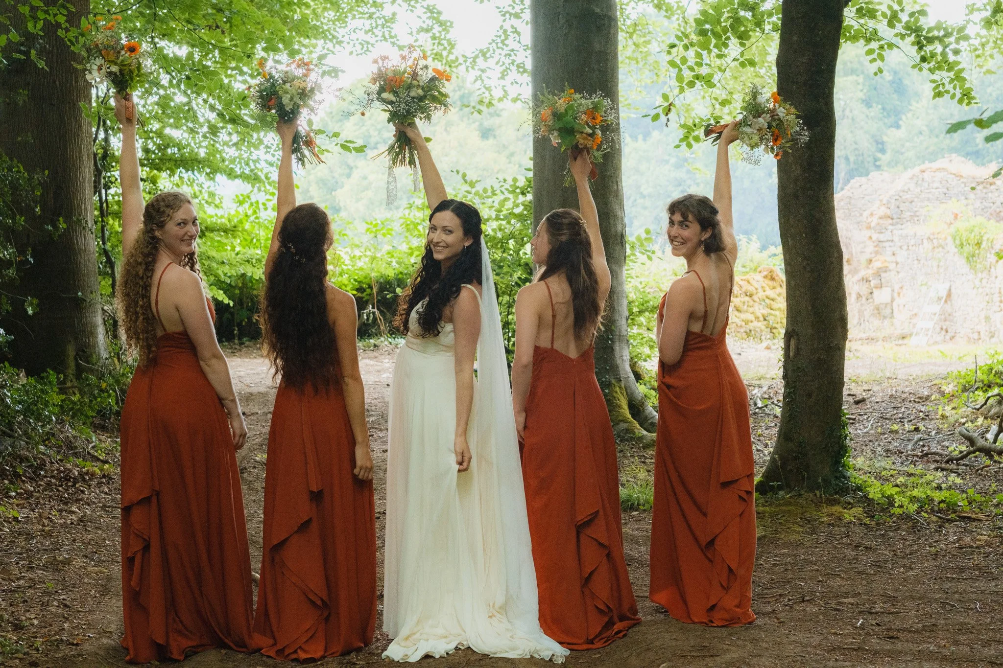 A bride with long dark curly hair in a white dress standing in a forest with five bridesmaids in rust-colored dresses, holding bouquets above their heads.