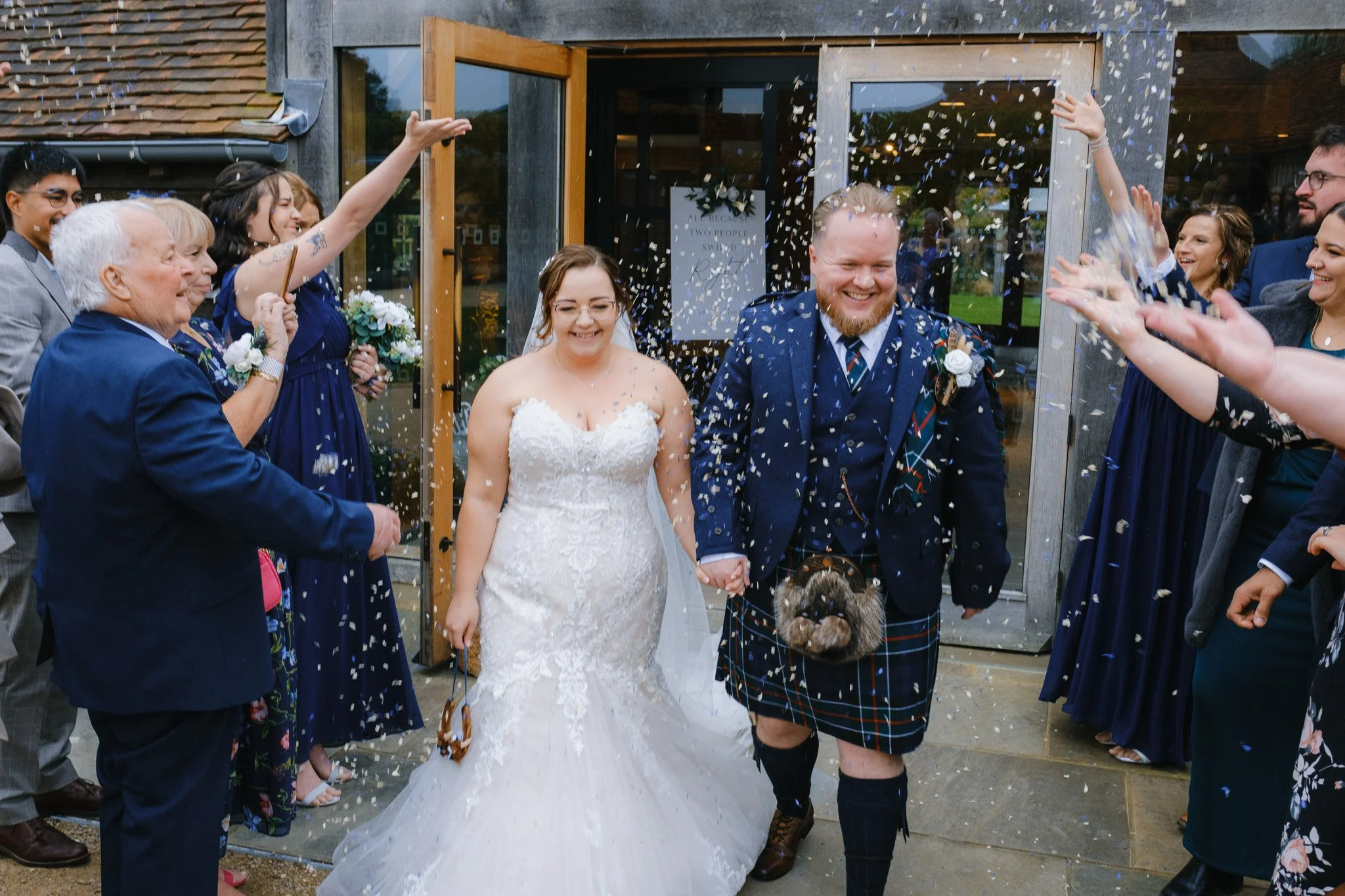 Bride and groom in wedding attire walk hand in hand while guests throw confetti outside a venue.