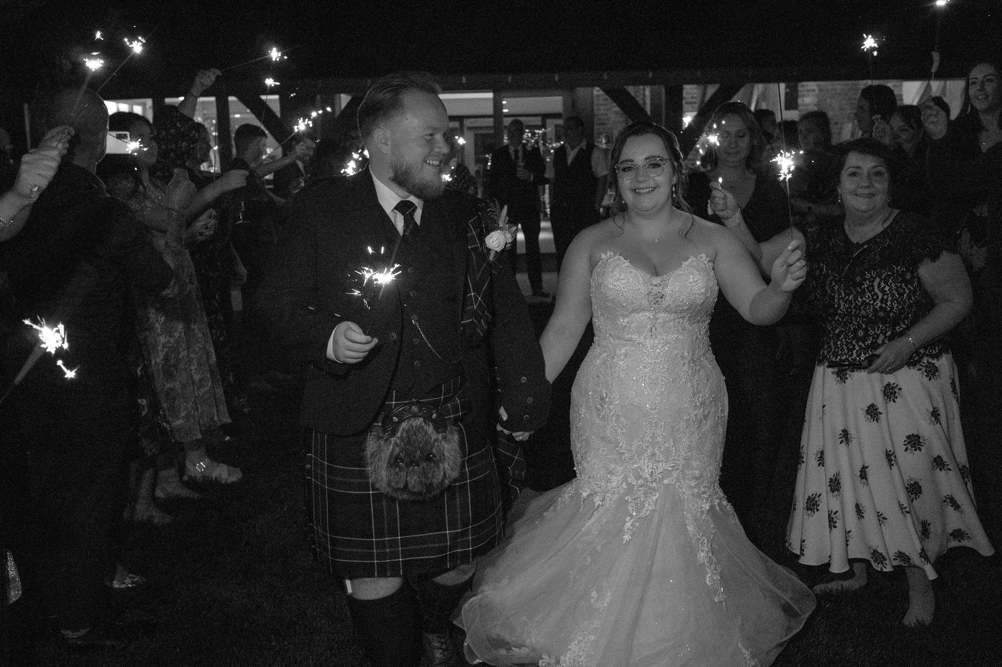 A black and white photo of a wedding reception with a bride in a lace gown and a groom in a kilt walking hand in hand. They are holding sparklers, surrounded by guests also holding sparklers. It's nighttime, and everyone looks joyful.