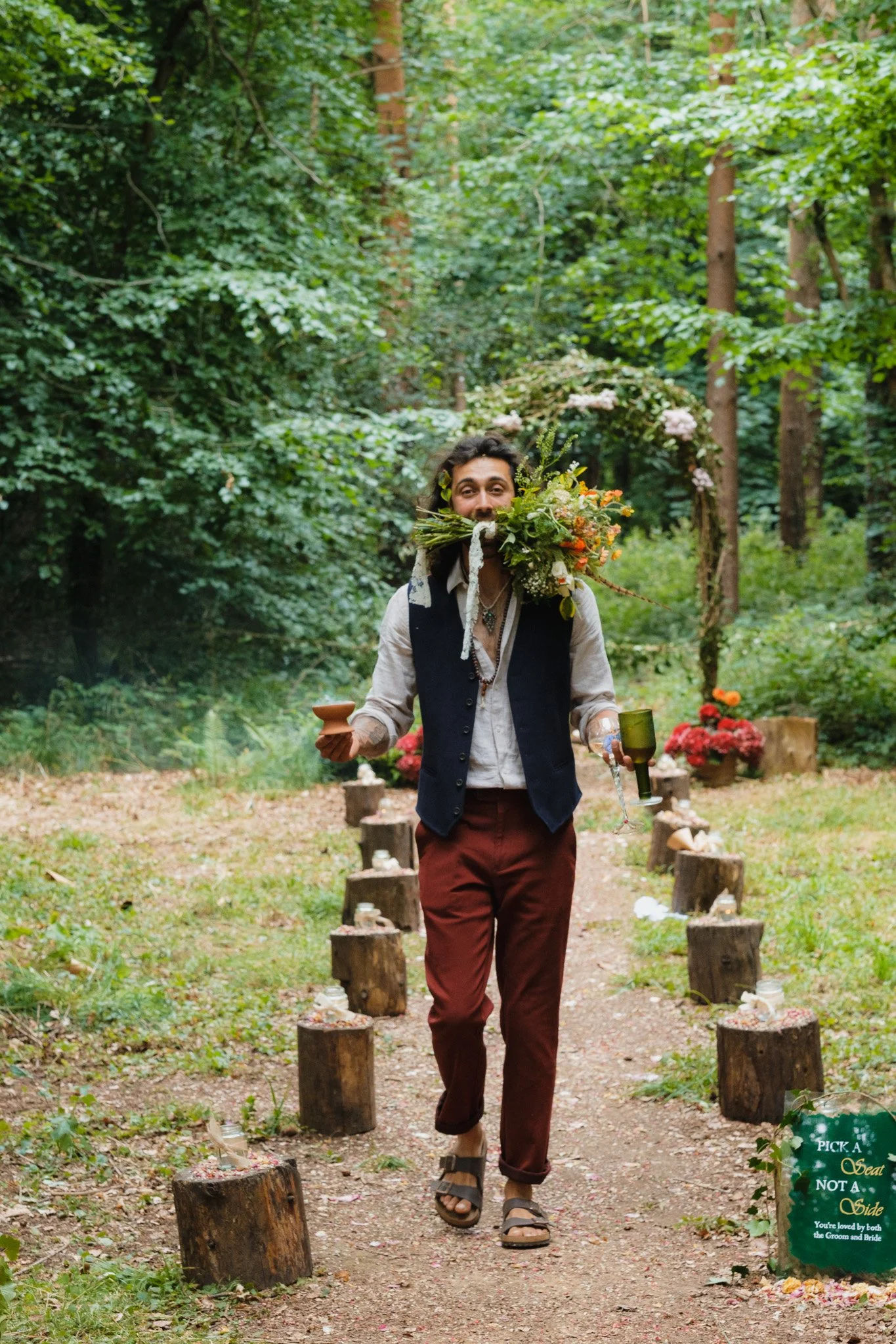 Man walking on a woodland trail holding a bouquet of flowers and two glasses, one in each hand, with a wedding setup in the background and a sign that reads 'Pick a Seat Not a Side'.