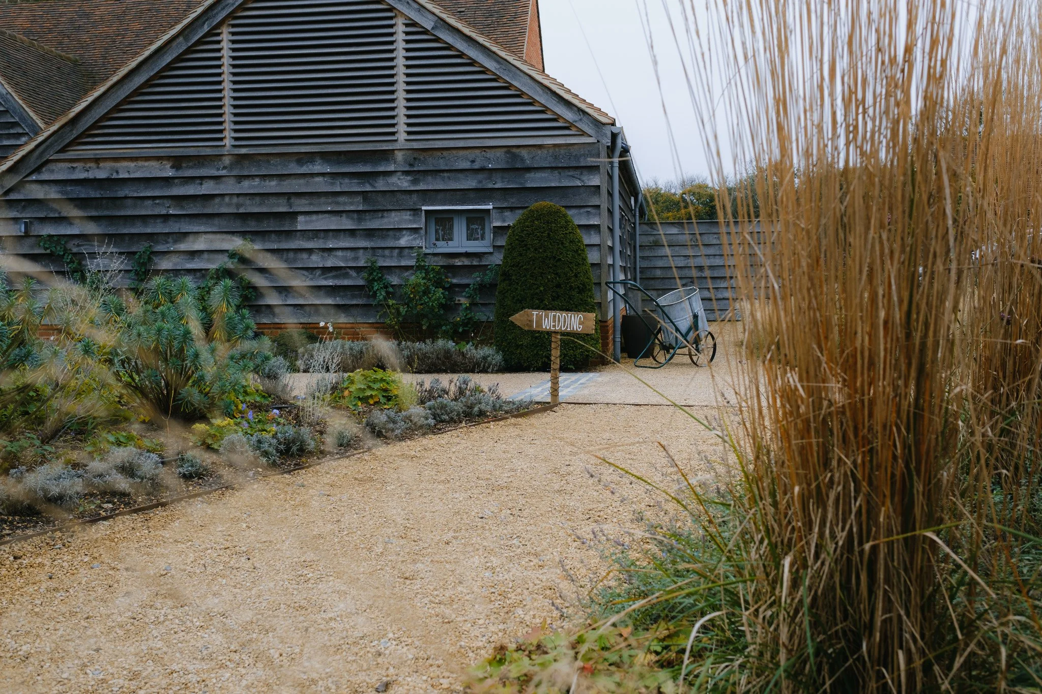 A rustic building with wooden siding and a garden path, featuring a sign reading "Wedding" in a landscaped yard.