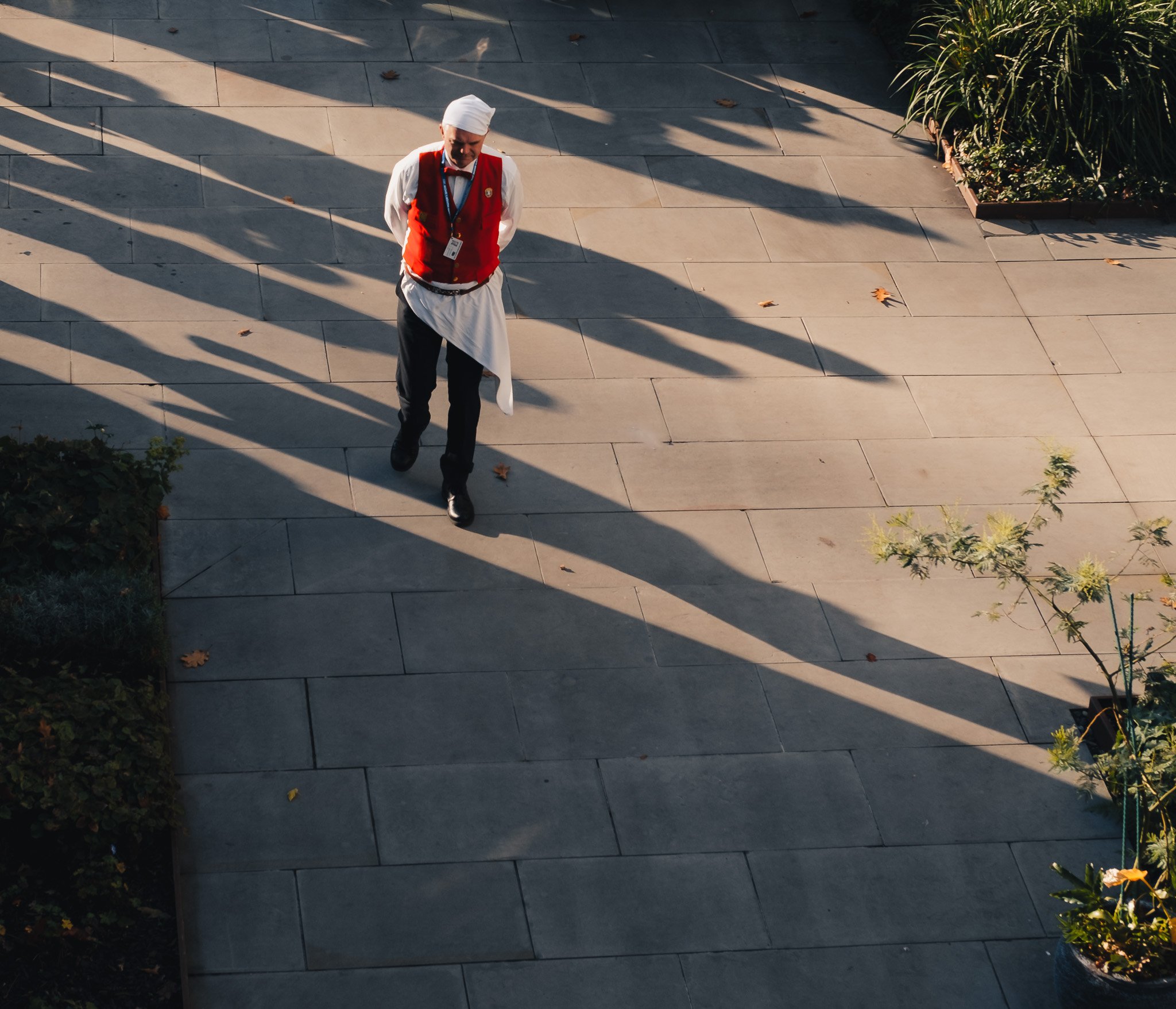 A person in a red vest and white headscarf walking on a paved path, casting long shadows. The path is surrounded by greenery and lit by sunlight.