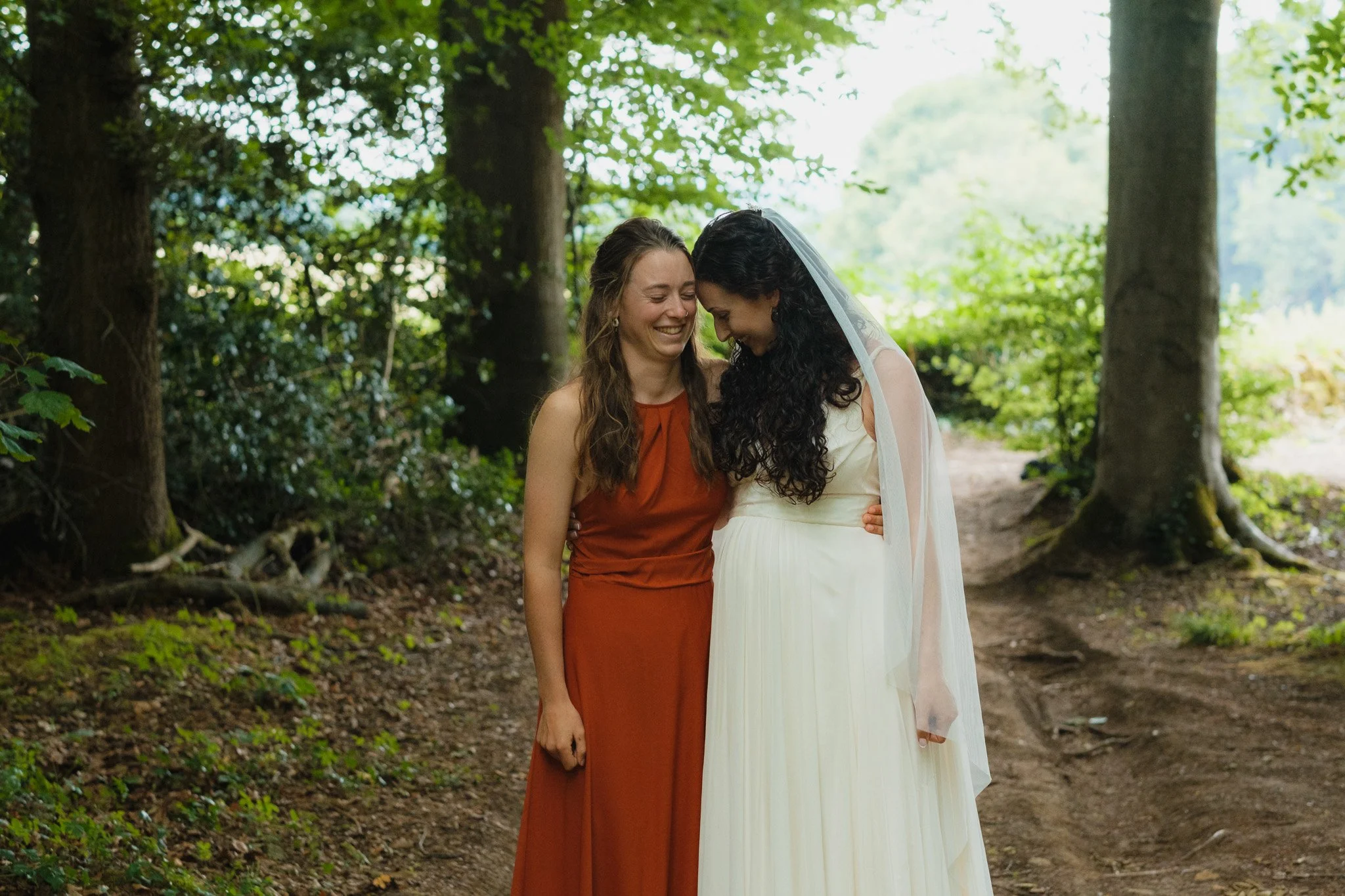 Two women in wedding attire, smiling and touching foreheads in a wooded outdoor setting.