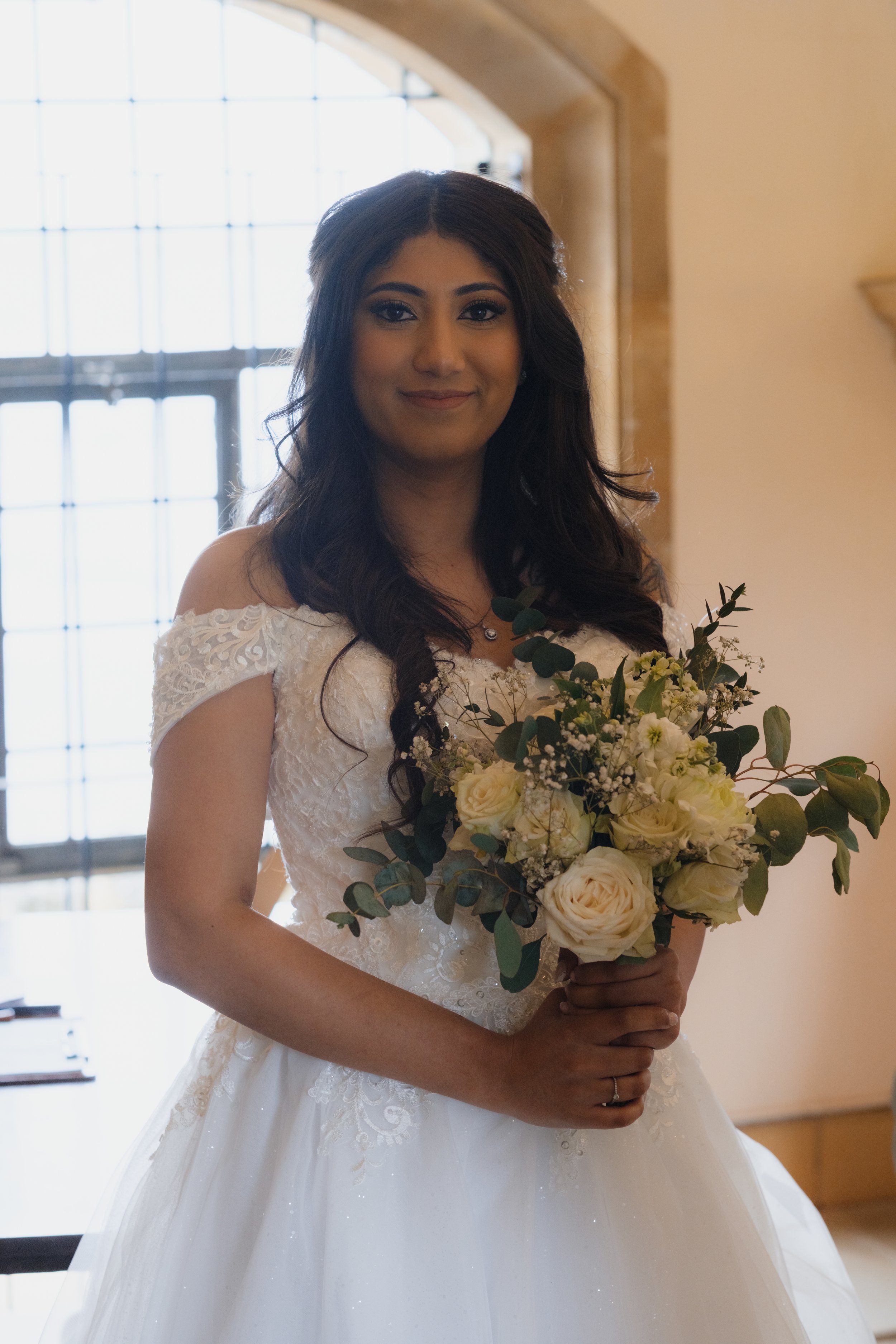 Bride in a white lace wedding dress holding a bouquet of white and green flowers indoors.