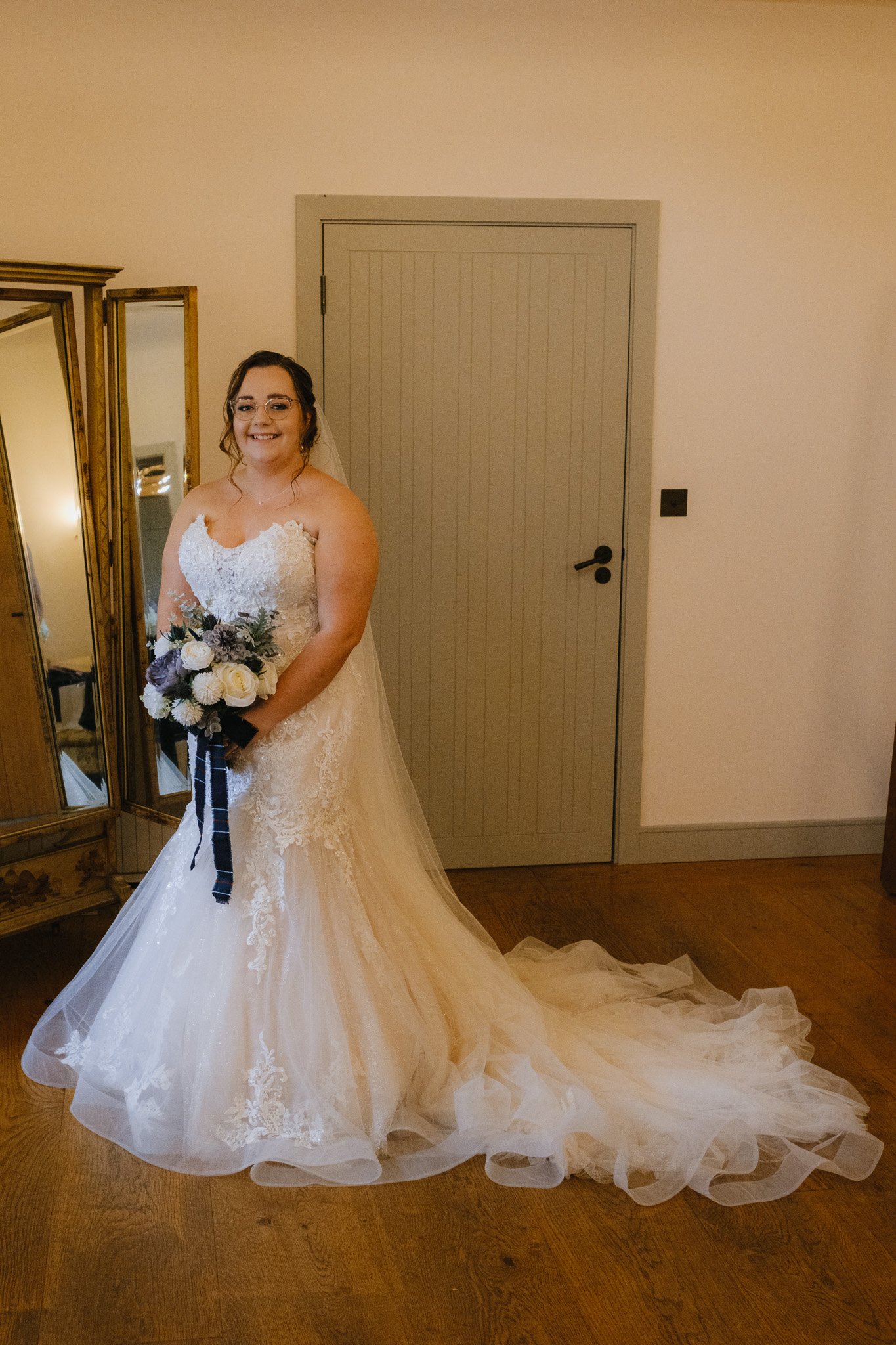 Bride in white lace wedding dress holding bouquet indoors with wooden floor and mirror.