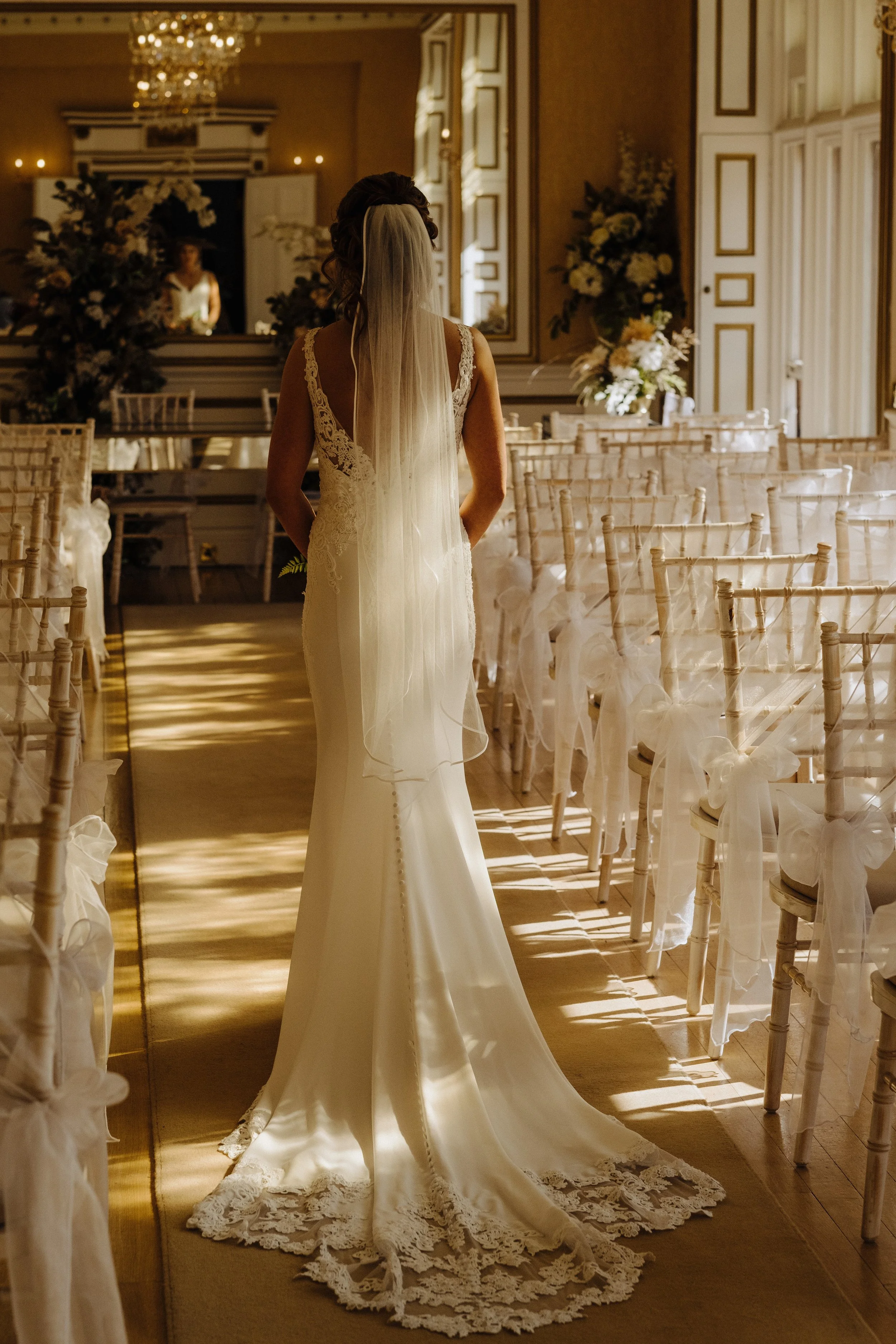 Bride in white dress walking down aisle at indoor wedding ceremony venue with chiavari chairs and floral decorations.