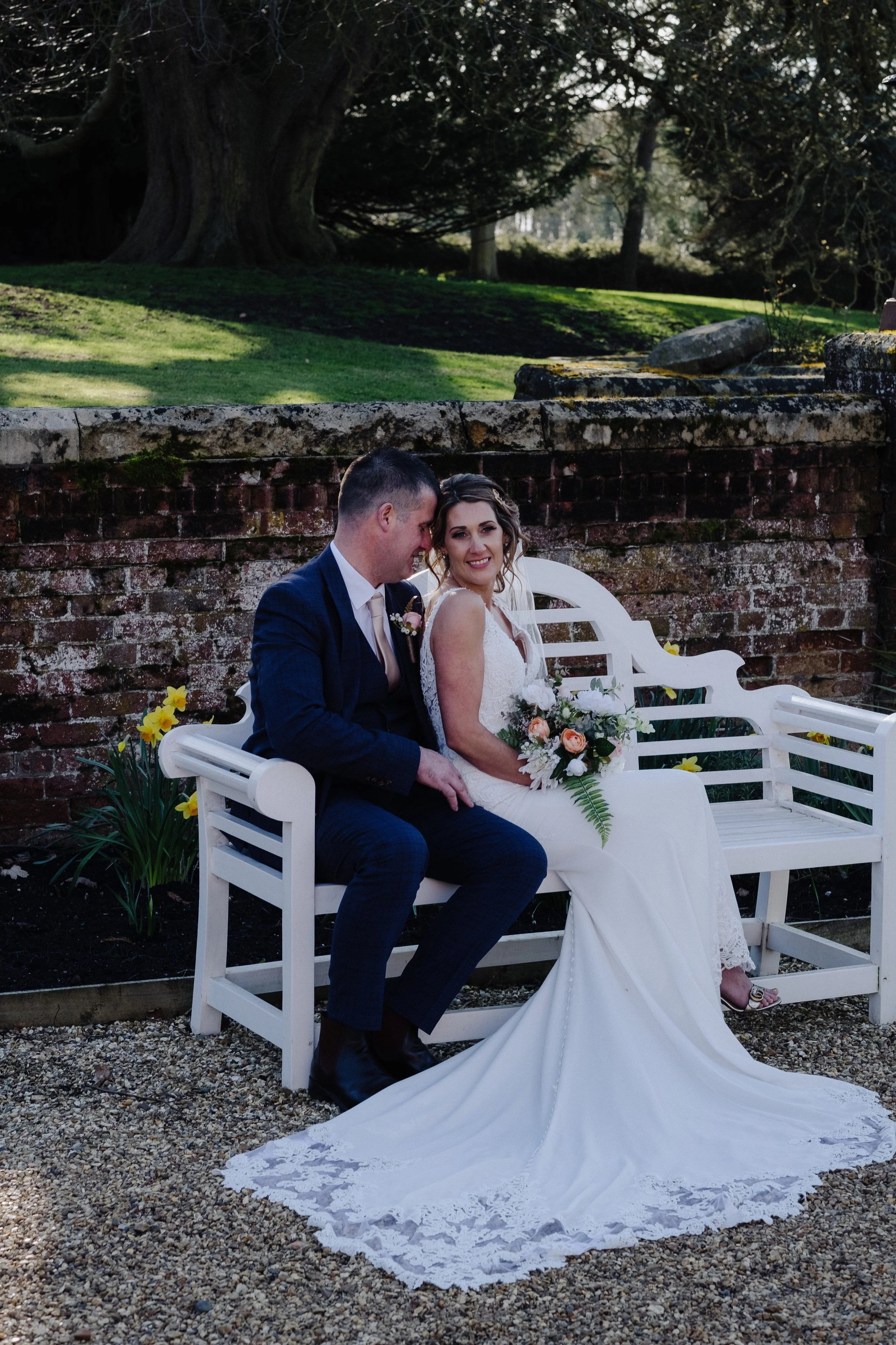 Bride and groom sitting on a white bench in a garden setting, with the bride in a wedding dress holding a bouquet.