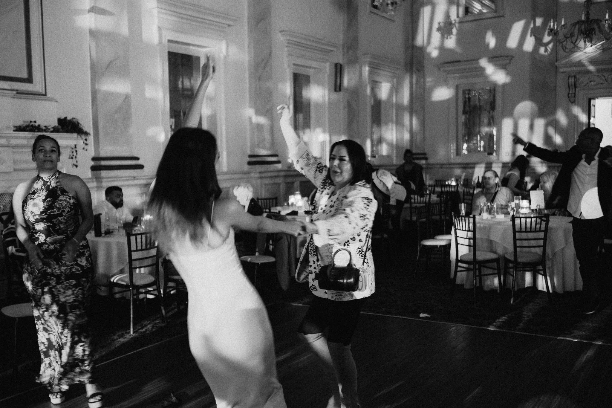 Black and white photo of people dancing in a formal indoor event, with tables and chairs in the background.