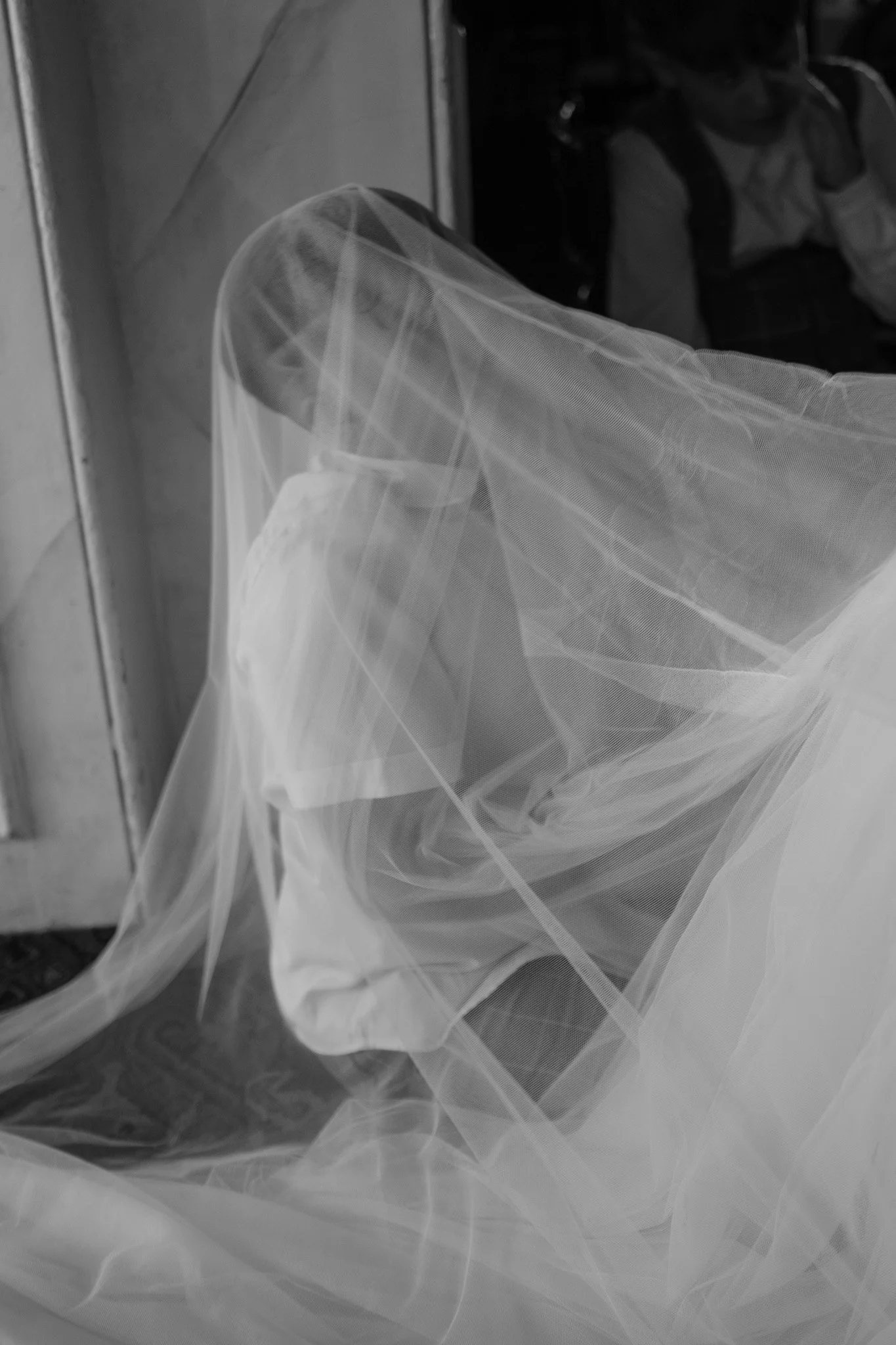 Black and white image of a person draped in sheer fabric, partially obscured, with another person in the background.
