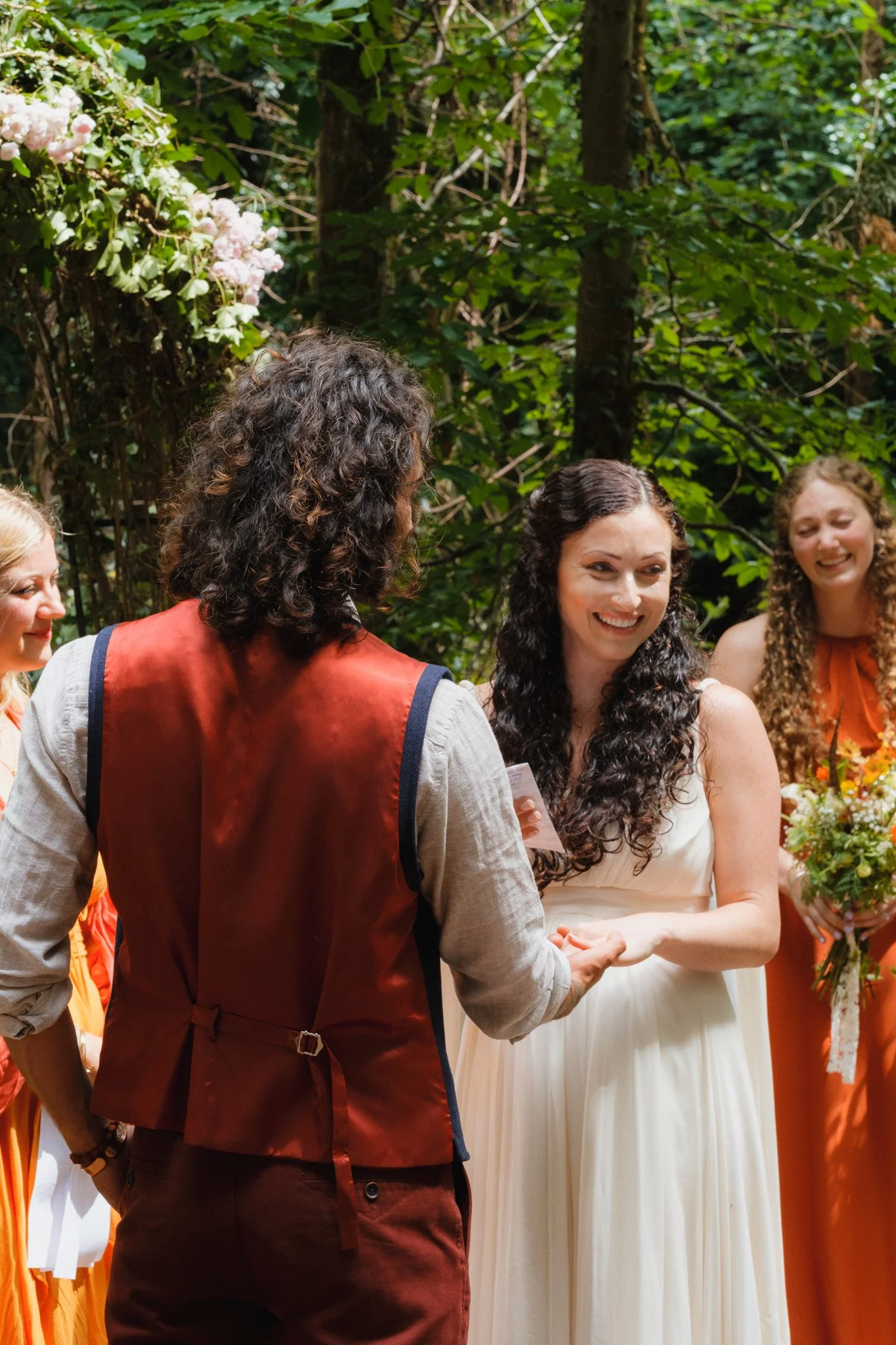A wedding ceremony outdoors with the bride and groom exchanging vows, surrounded by bridesmaids holding bouquets, and lush green trees in the background.
